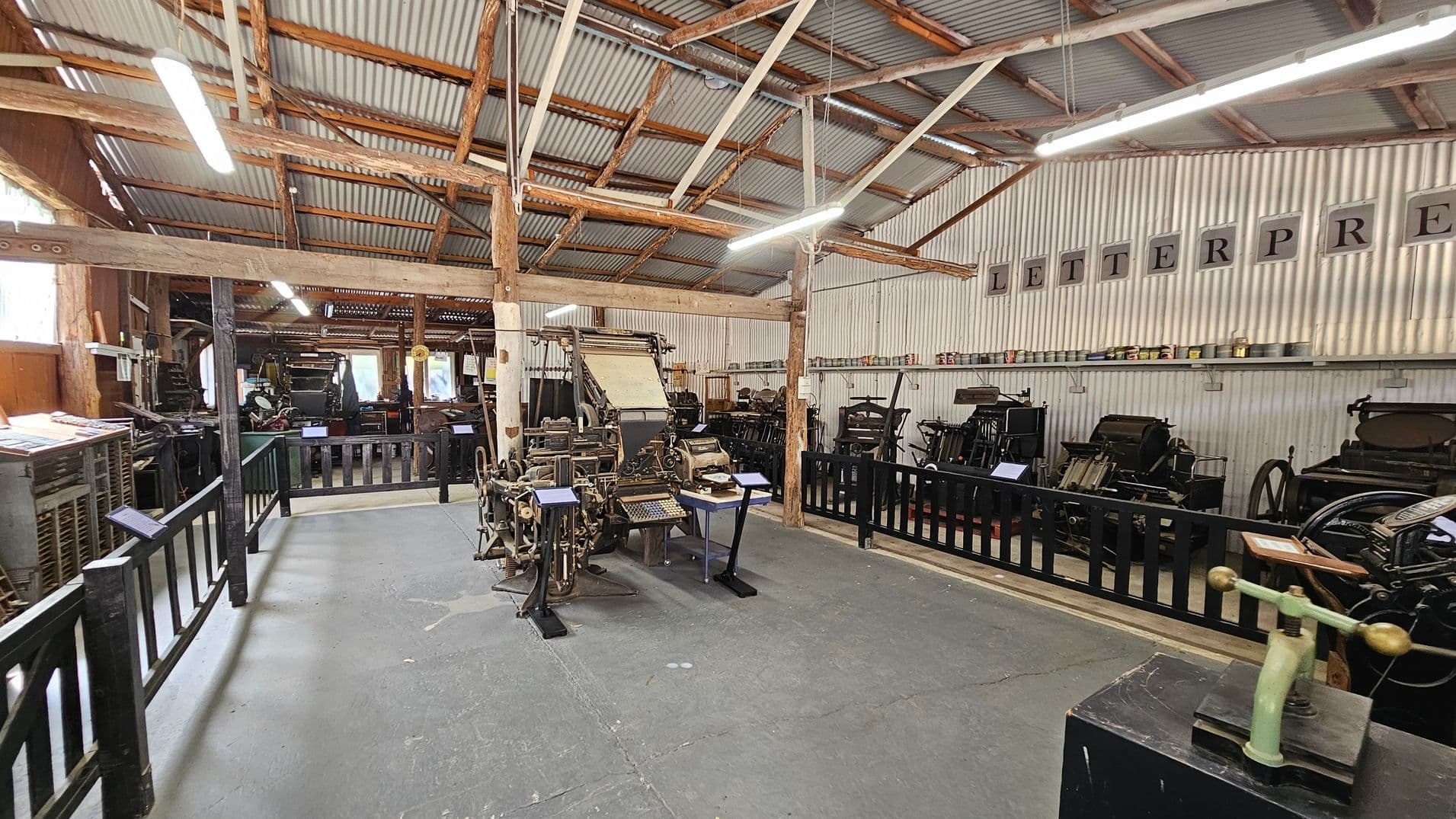 A room with a corrugated metal ceiling and wooden beams. On the left, there are various vintage printing machines behind a black wooden fence. In the centre, a large, intricate printing press is displayed with informational plaques. On the right, more printing equipment is lined up against the wall, which has the word "LETTERPRESS" displayed in large letters. The floor is concrete, and fluorescent lights hang from the ceiling.
