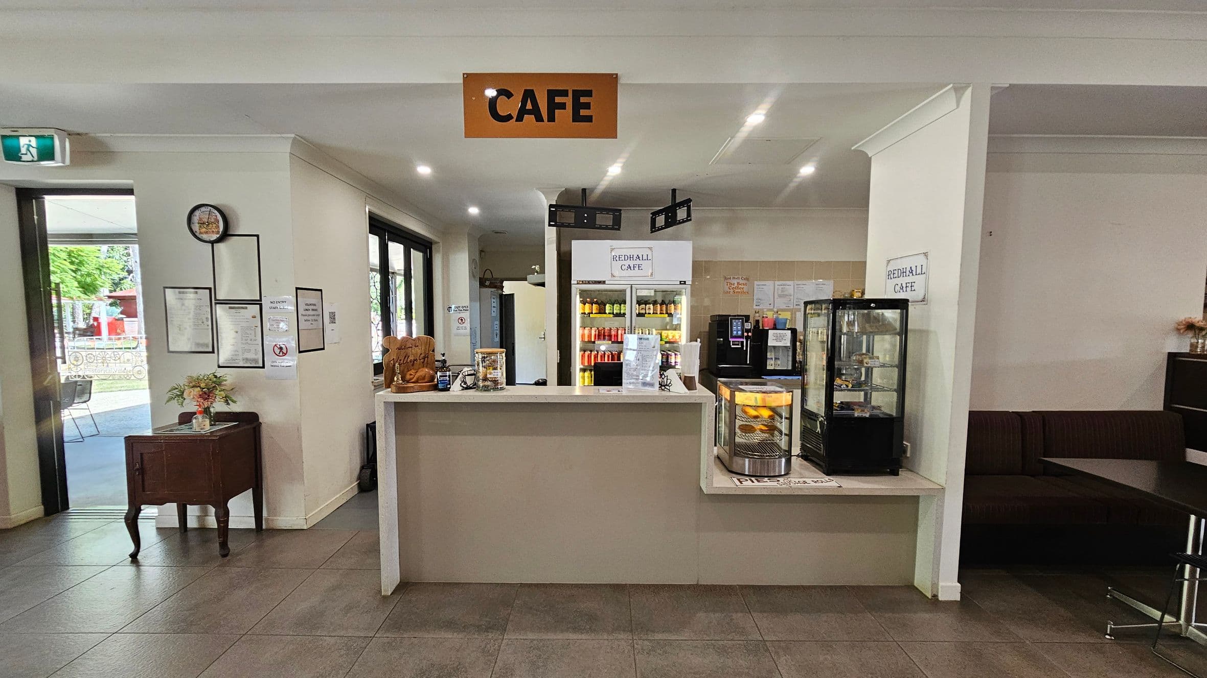 A café interior with a central counter displaying a small glass case containing pastries. Behind the counter, a fridge is stocked with various drinks. To the left, a small table with flowers and framed notices on the wall. An exit sign and glass doors lead outside. On the right, a seating area with a dark bench. The ceiling is white with recessed lighting, and the floor is tiled in a greyish tone. A sign above reads "CAFE".