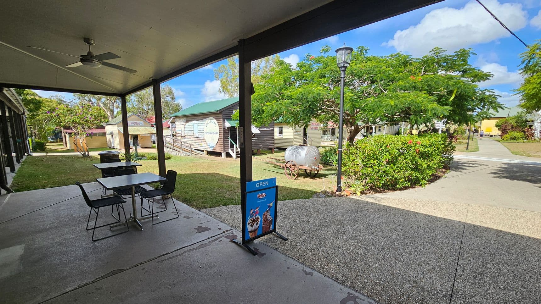A covered outdoor area with a ceiling fan and two tables with chairs on a concrete floor. A sign with ice cream images and the word "OPEN" is in the foreground. In the background, there are wooden buildings with green roofs, one with a "Welcome" sign. A vintage water cart is near a tree with lush green foliage. A lamppost stands beside a path leading through a garden with bushes and flowers.