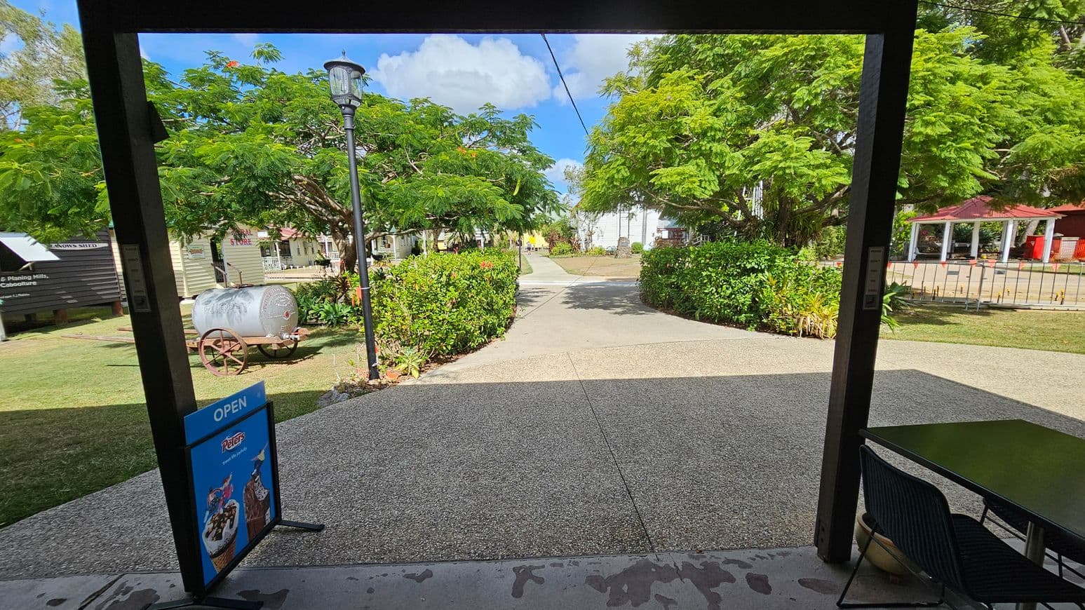 A view from a shaded area with a sign reading "OPEN" featuring ice cream images on the left. A table and chair are on the right. Beyond, a path leads through a garden with lush green trees and bushes. A vintage metal cart is on the left, and a lamppost stands nearby. In the background, there are small buildings and a red-roofed structure. The sky is partly cloudy.