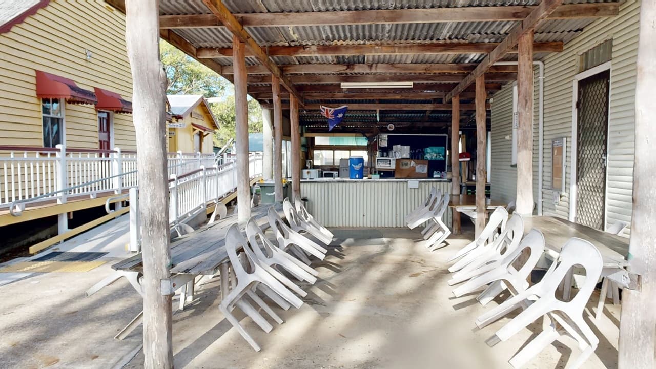 A covered outdoor area with a corrugated metal roof supported by wooden beams. In the centre, a bar or serving counter is visible, with various items like a water cooler and microwave. Plastic chairs are stacked on both sides, leaning against wooden tables. To the left, a wooden building with red awnings and a white railing is visible. The floor is concrete, and the ceiling is made of corrugated metal. Trees can be seen in the background.