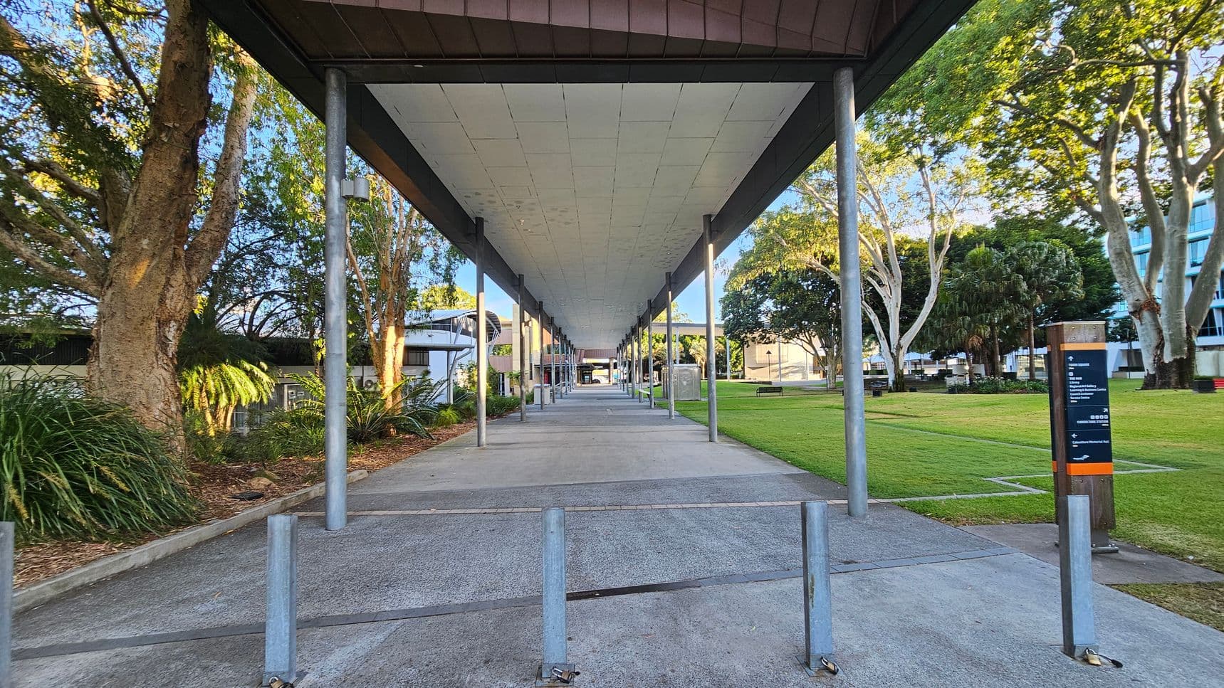A covered walkway with a series of metal poles runs through the centre, leading to a distant building. On the left, there are large trees and dense greenery. On the right, a grassy area with more trees and a signpost. The ground is a mix of concrete and paved surfaces. The ceiling of the walkway is flat and light-coloured.