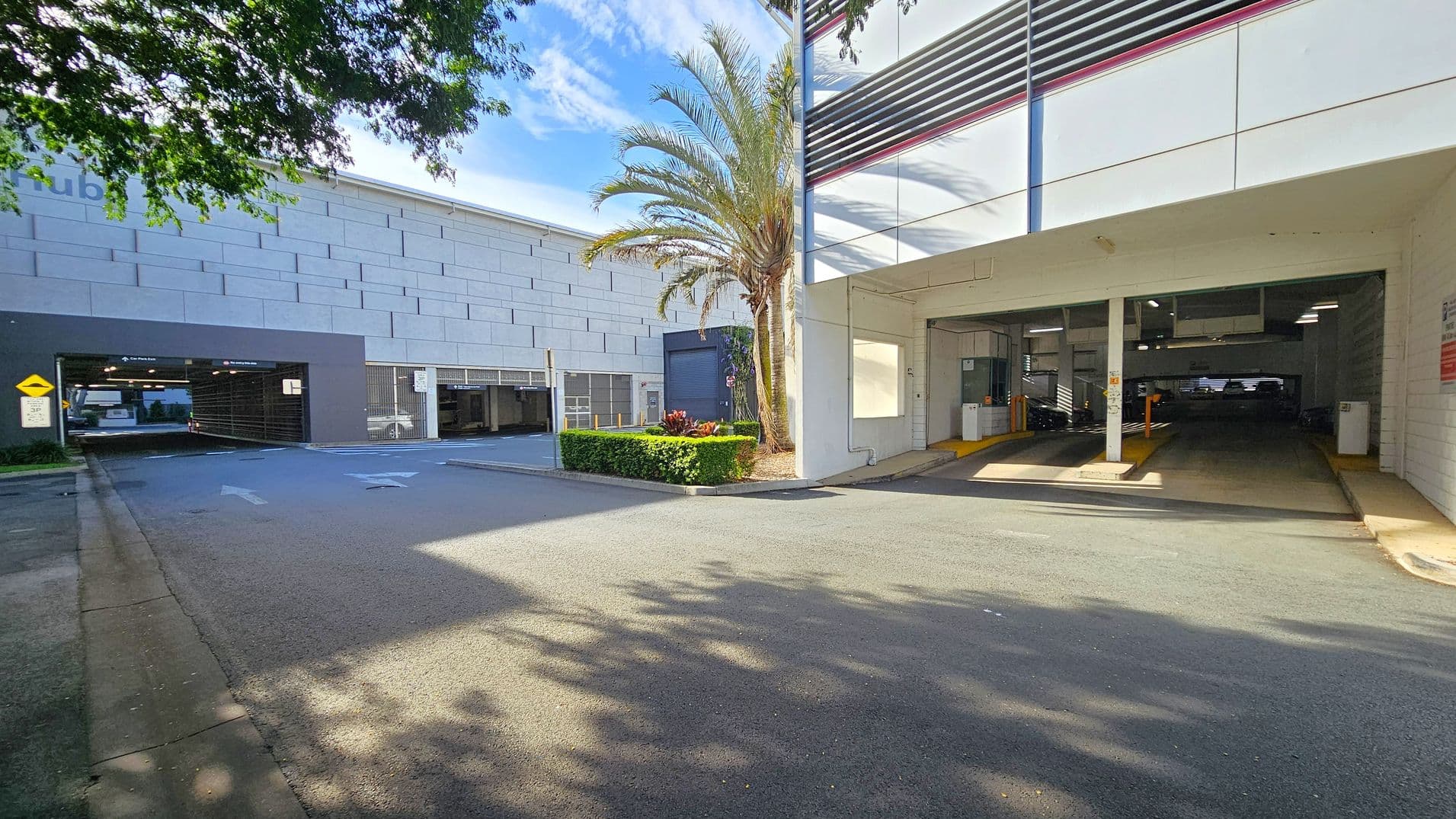 A parking area with two entrances. On the left, a multi-storey car park with a grey facade and an entrance marked by a yellow sign. In the centre, a palm tree and some shrubs are planted in a small landscaped area. On the right, another entrance to a covered parking area with white walls and a striped design above. The ground is paved, and there are shadows from nearby trees.