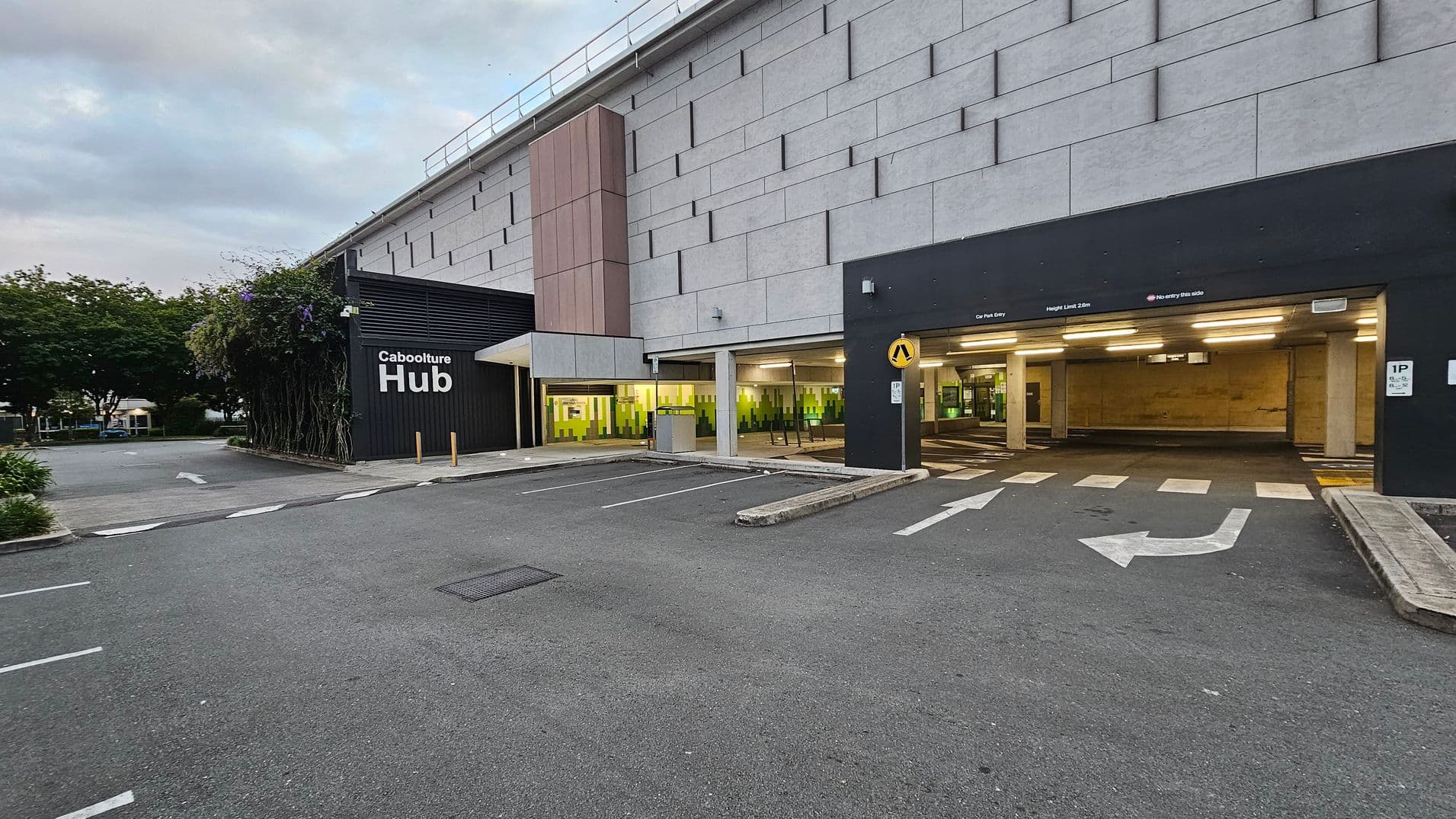 A large building with a textured facade and a tall vertical panel in the centre. On the left, there's a sign reading "Caboolture Hub" next to a wall covered with climbing plants. The entrance to a car park is on the right, marked by a black overhead sign and illuminated interior. The ground is paved with directional arrows and parking spaces. Trees and greenery are visible in the background on the left.