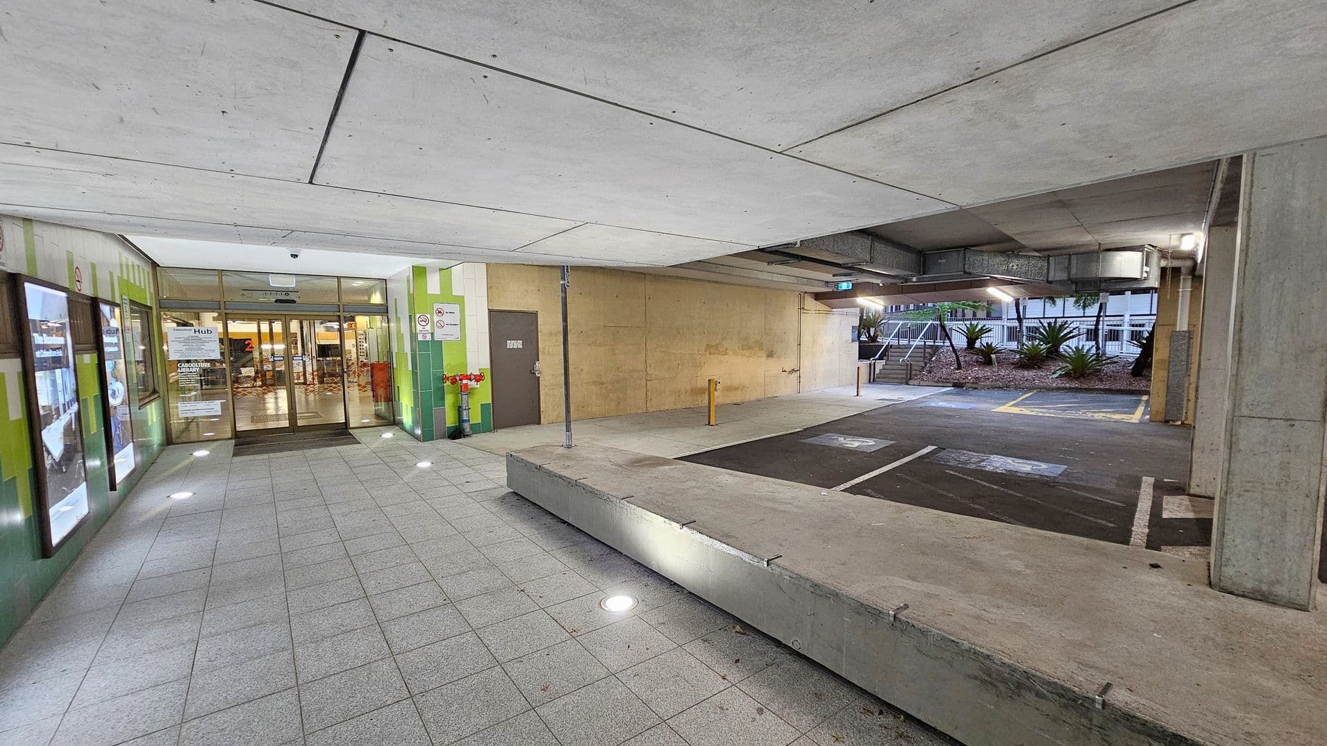 A covered entrance area with a tiled floor and a concrete ceiling. On the left, there are glass doors leading into a building, surrounded by green and beige walls with signage. Illuminated display panels are mounted on the wall. On the right, there is a parking area with marked spaces and a few bollards. In the background, there are plants and a building with windows. Ceiling lights are embedded in the floor, providing illumination.