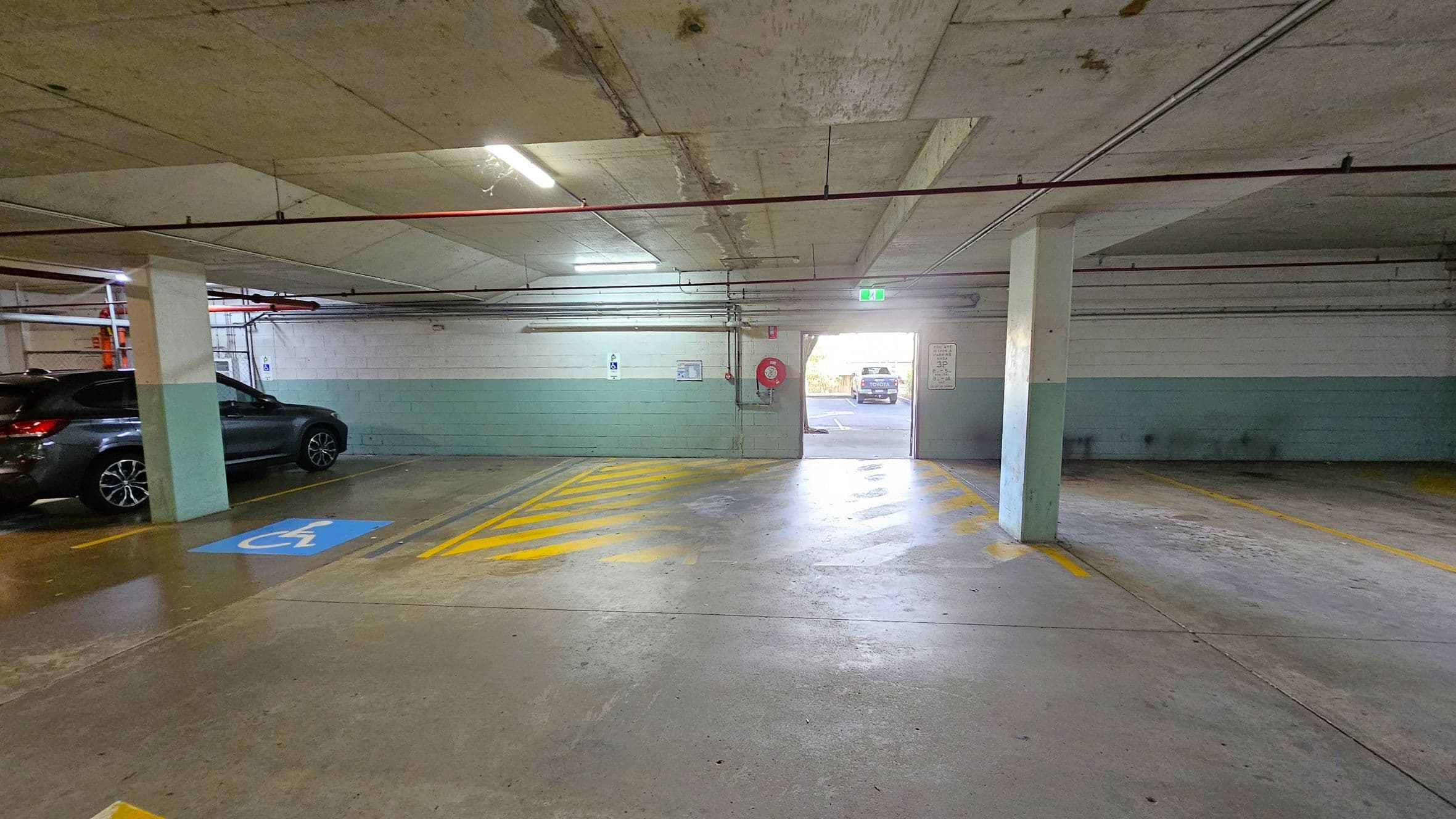 A parking garage with a concrete floor and ceiling. On the left, a dark-coloured car is parked next to a column. A blue disabled parking symbol is painted on the ground. In the centre, yellow diagonal lines mark a no-parking zone leading to an exit. The exit shows daylight and a vehicle outside. On the right, empty parking spaces are visible. Red pipes run along the ceiling, and a fire hose reel is mounted on the back wall.