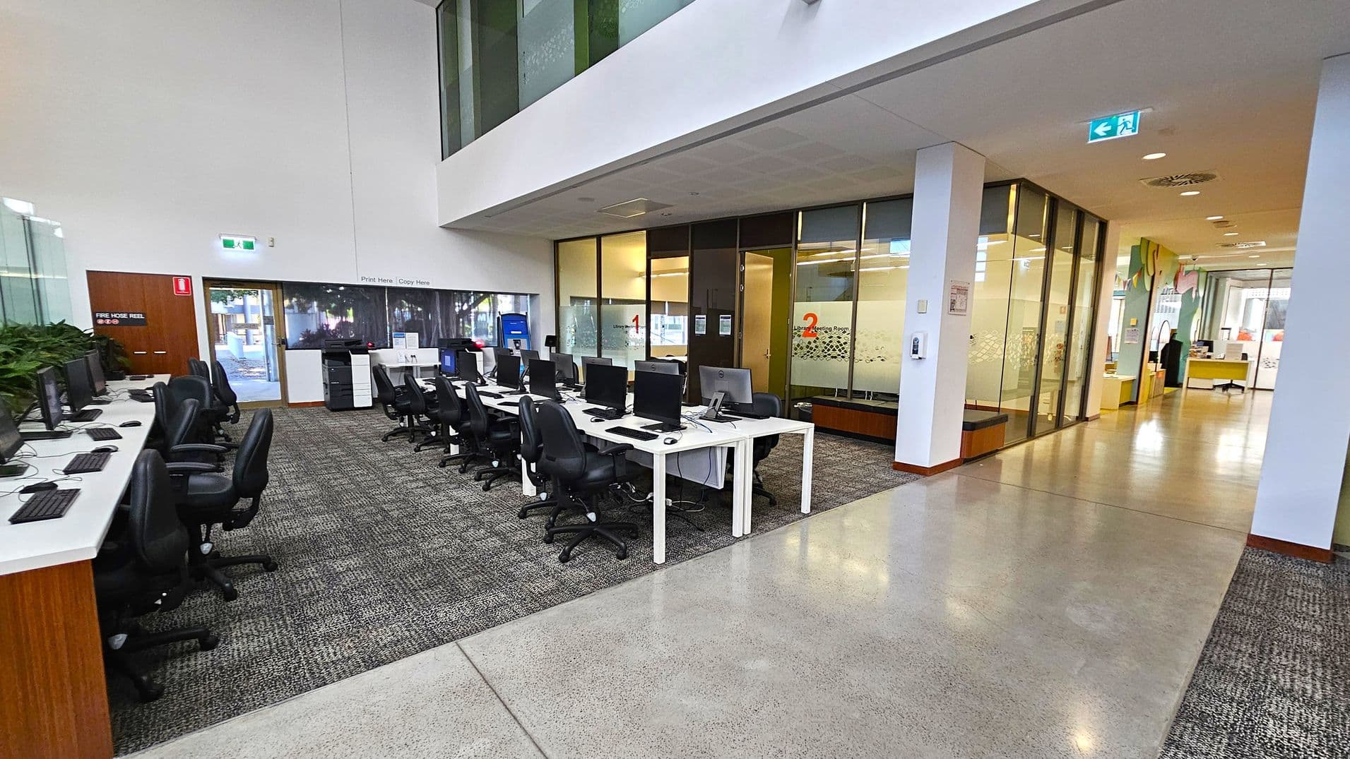 On the left, there are rows of computer desks with black chairs on a carpeted floor. A fire hose reel is mounted on the wall near a door. In the centre, there are more computer stations and a printer area. On the right, glass-walled meeting rooms are visible, labelled with numbers. The corridor leads to an open area with more office furniture. The ceiling is white with recessed lighting, and the floor transitions from carpet to polished concrete.
