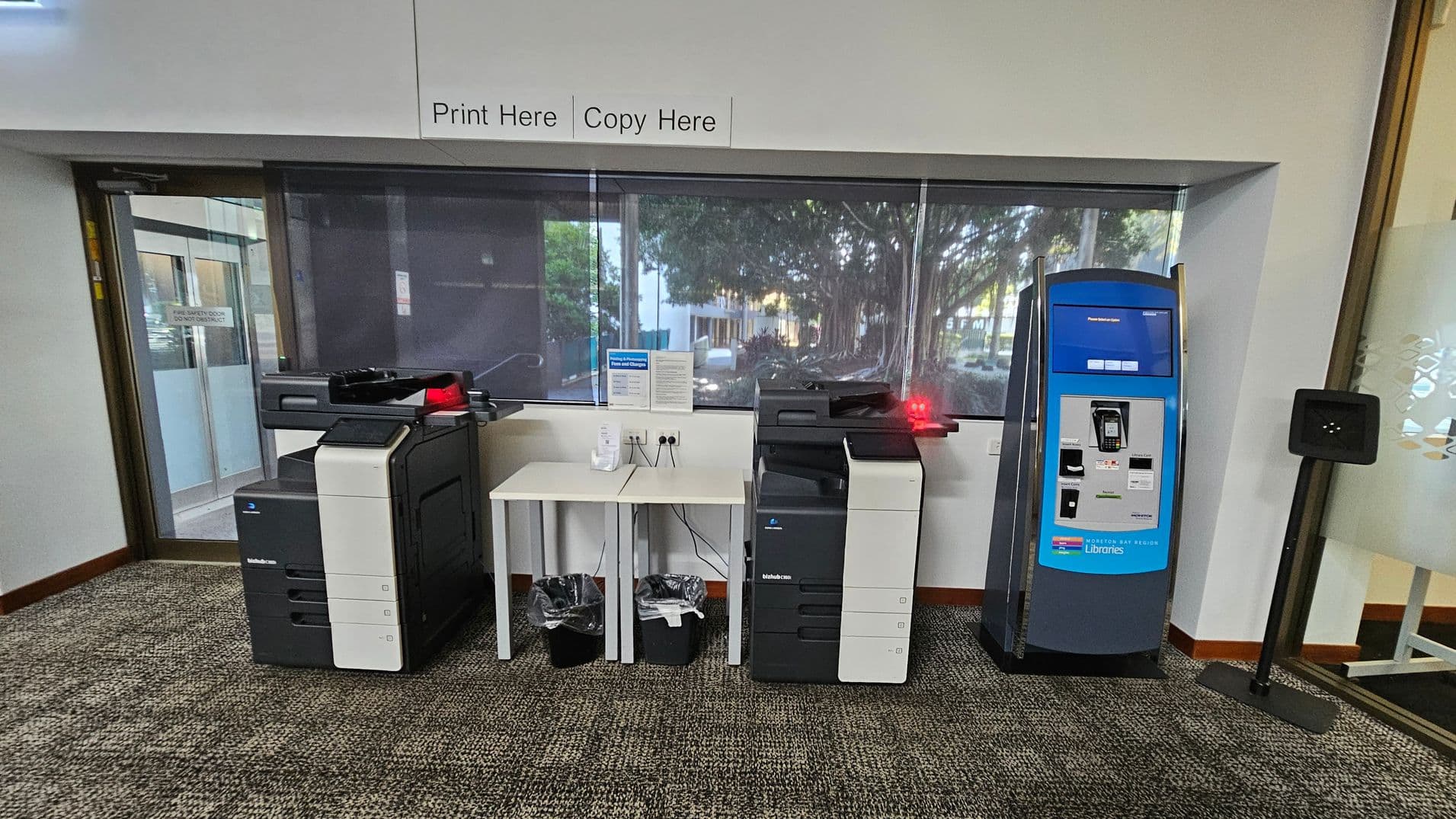 On the left, there is a large printer with a black and white design. In the centre, a small white table holds a hand sanitiser dispenser, with two bins underneath. To the right, another similar printer is positioned next to a blue payment kiosk. The floor is carpeted with a patterned design, and a large window in the background shows trees outside. A sign above reads "Print Here Copy Here".