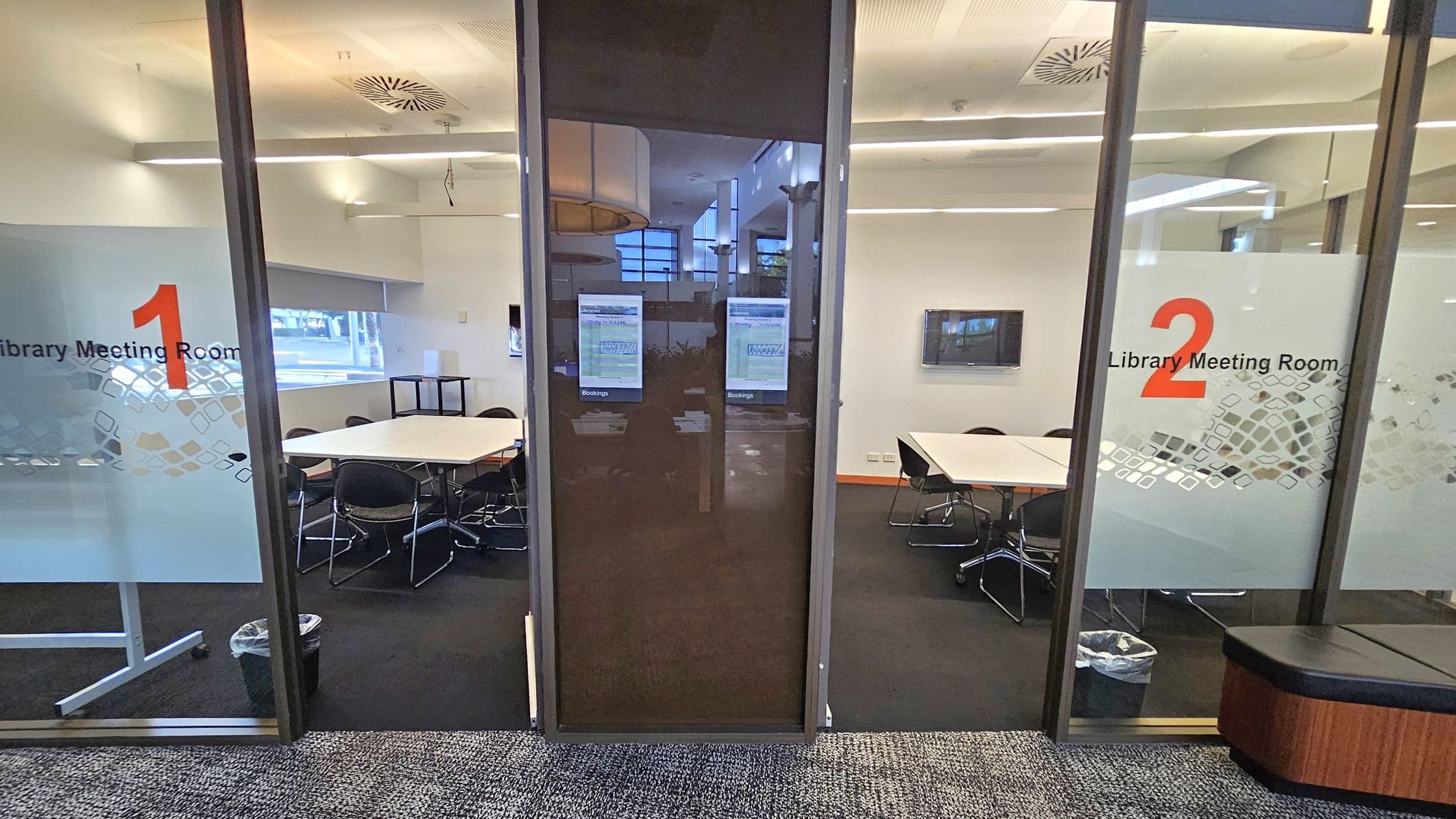 A view of two library meeting rooms separated by a central glass door. On the left, Meeting Room 1 has a white table surrounded by black chairs and a small bin. On the right, Meeting Room 2 features a similar setup with a wall-mounted screen. Both rooms have frosted glass panels with numbers and the words "Library Meeting Room." The floor is carpeted, and the ceiling has recessed lighting and ventilation panels.