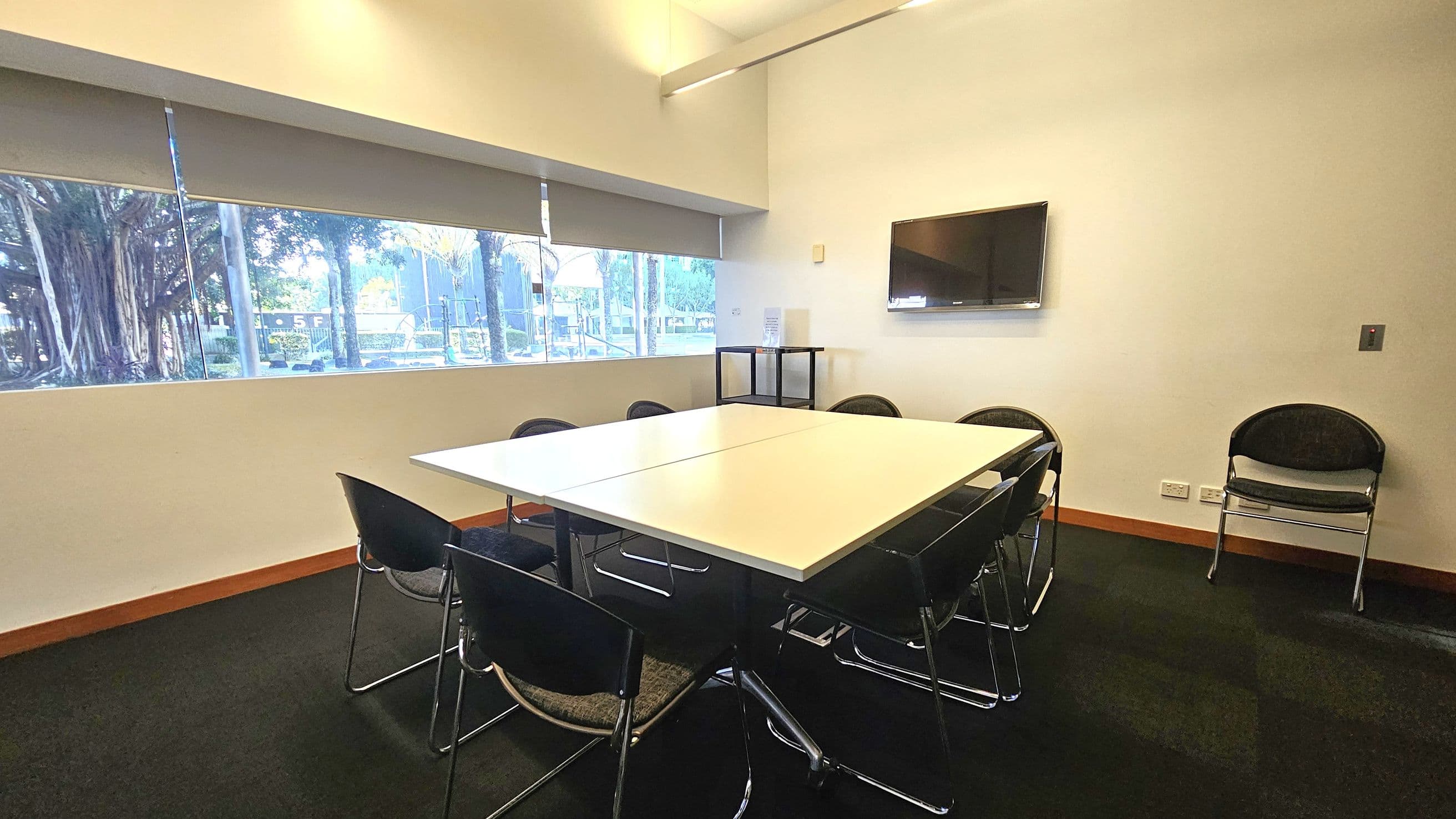A meeting room with a large white table in the centre surrounded by black chairs. On the right wall, a flat-screen TV is mounted above a small black shelf. The left side features large windows with blinds partially drawn, revealing trees and a building outside. The floor is carpeted in dark tones, and the walls are plain white. Additional chairs are stacked in the corner. The ceiling is not visible.