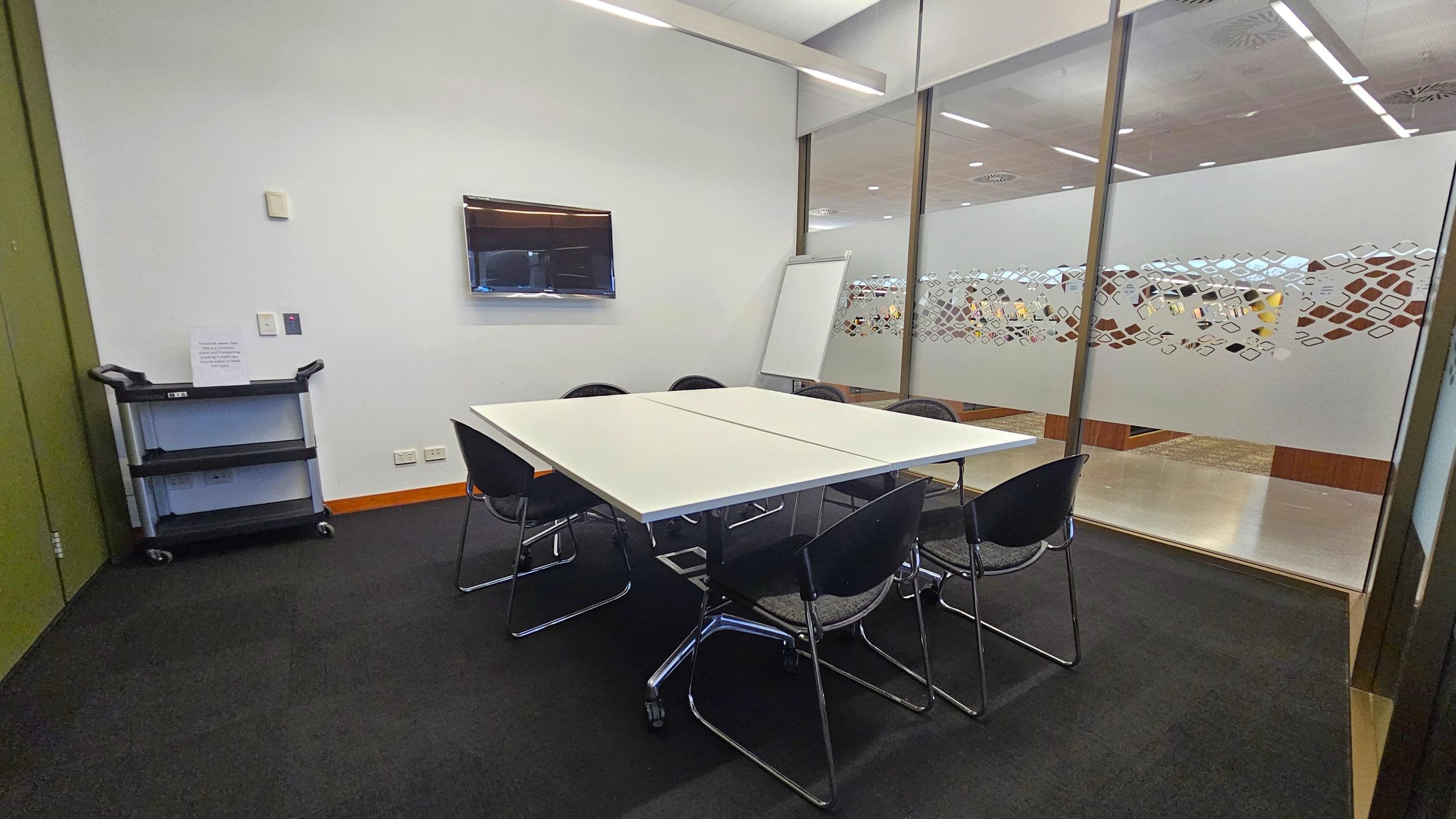 A small meeting room with a rectangular table in the centre surrounded by six black chairs. On the left, a black utility cart with shelves is against the wall. A flat-screen monitor is mounted on the wall above the cart. The right side features a glass partition with a decorative pattern, and a whiteboard is positioned nearby. The floor is carpeted in dark grey, and the ceiling has recessed lighting.