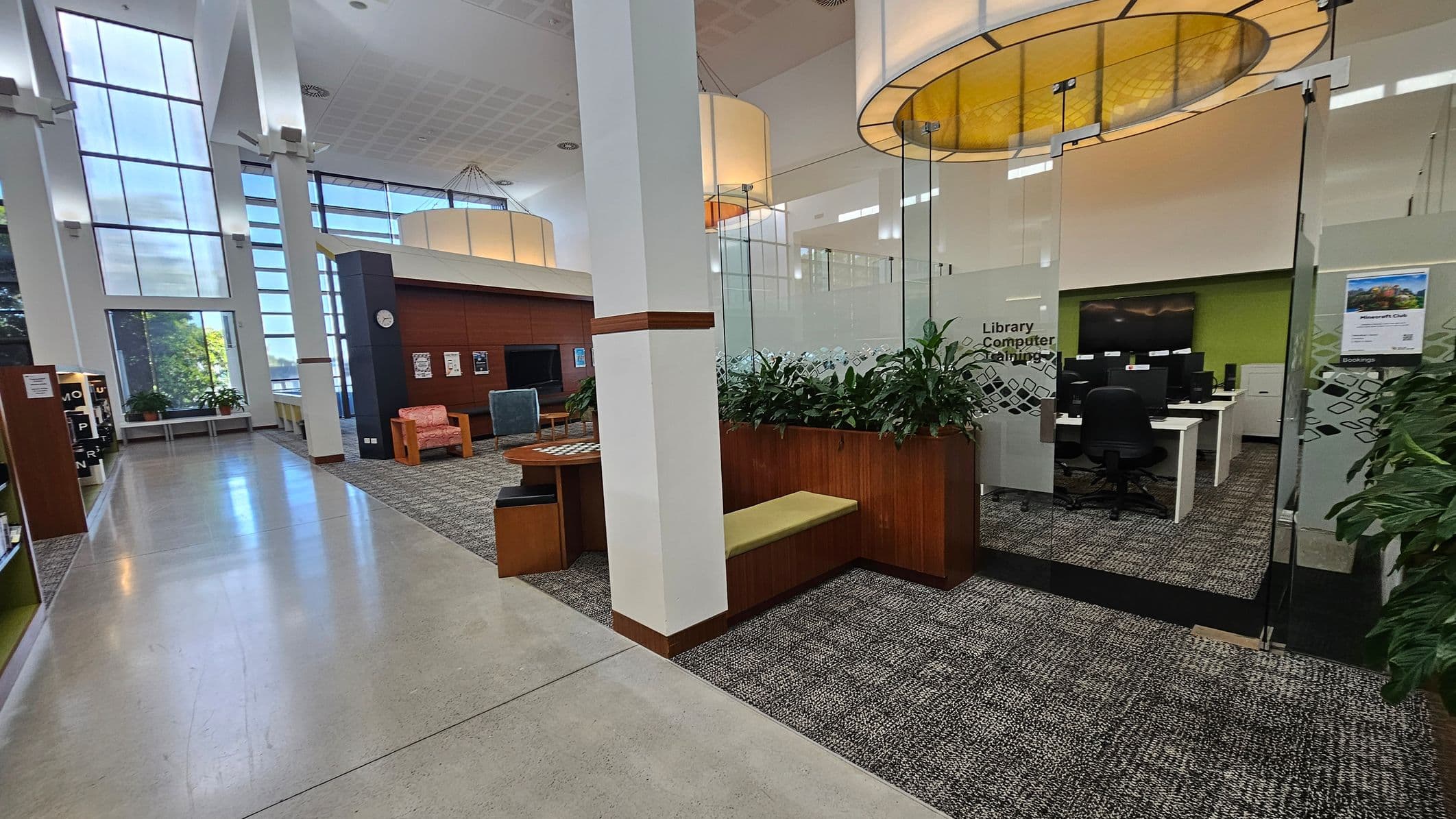 A spacious library interior with large windows on the left, allowing natural light to flood in. The floor is a mix of polished concrete and carpet. On the right, there's a glass-walled room labelled "Library Computer Catalogue" with several computer stations and chairs. In the foreground, a wooden bench with a cushion and a planter with green plants. The ceiling features large, circular light fixtures. In the background, there are bookshelves and seating areas with armchairs.