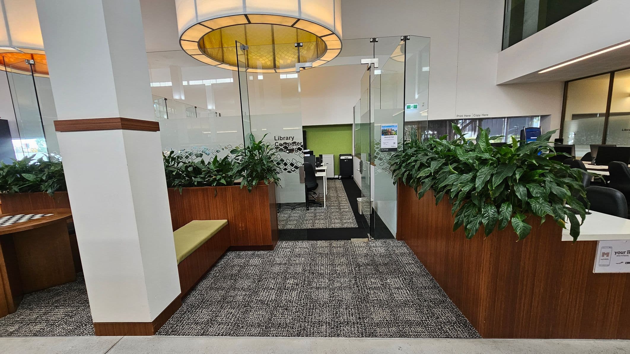 A modern library interior with a central entrance flanked by glass walls. On either side, there are wooden planters with lush green plants. A cushioned bench is on the left, and a row of computers is visible through the glass. The floor is carpeted with a patterned design, and a large circular light fixture hangs from the ceiling. The walls are white, and there are signs indicating library services.