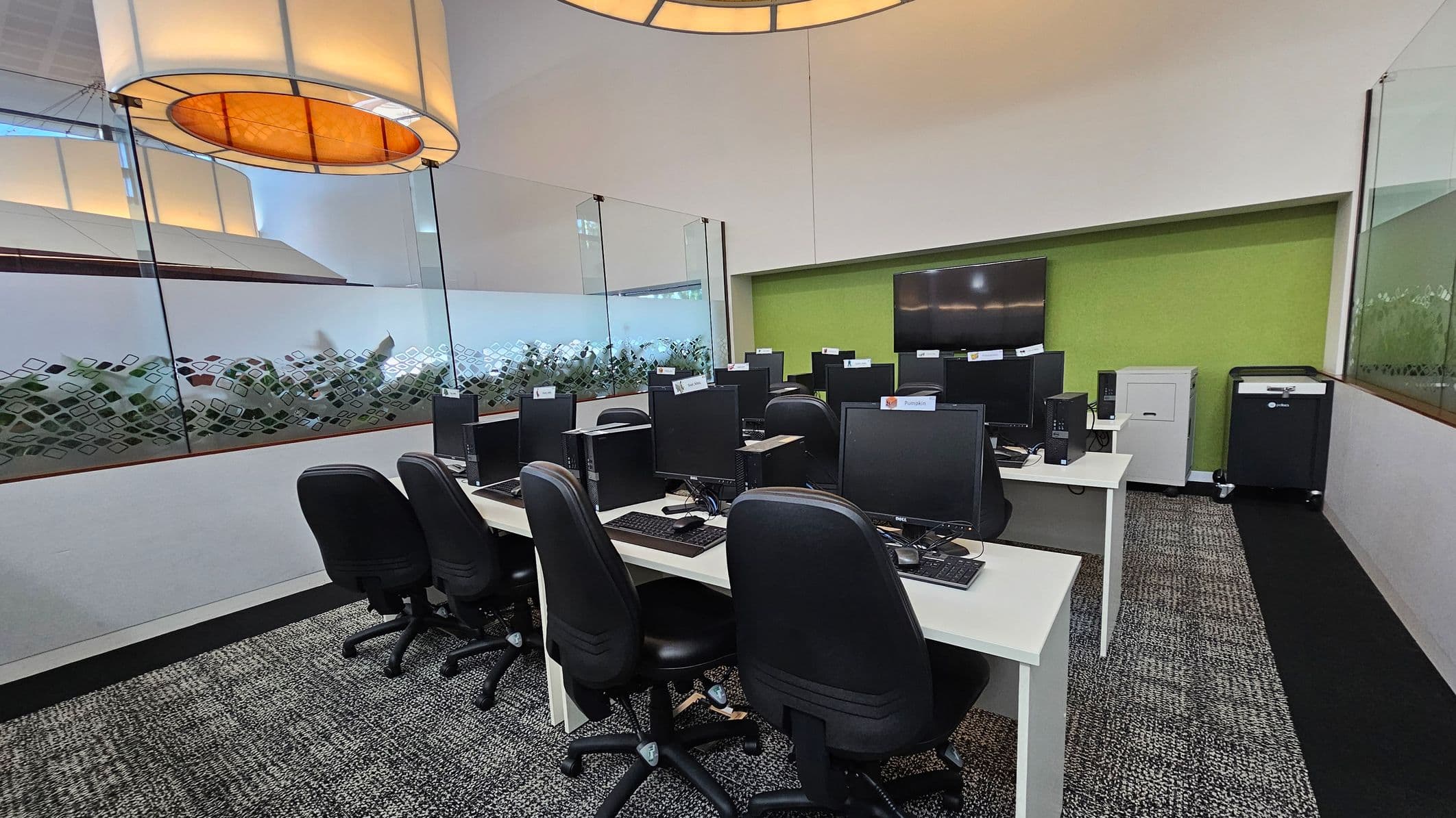 A modern computer lab with several black office chairs and computers arranged on white desks. Each computer has a name tag. The floor is carpeted in a grey pattern. The back wall is green with a large flat-screen TV mounted on it. To the right, there is a white cabinet and a black cart. The left and right walls have glass panels with a decorative pattern. Large circular ceiling lights hang above, casting a warm glow.