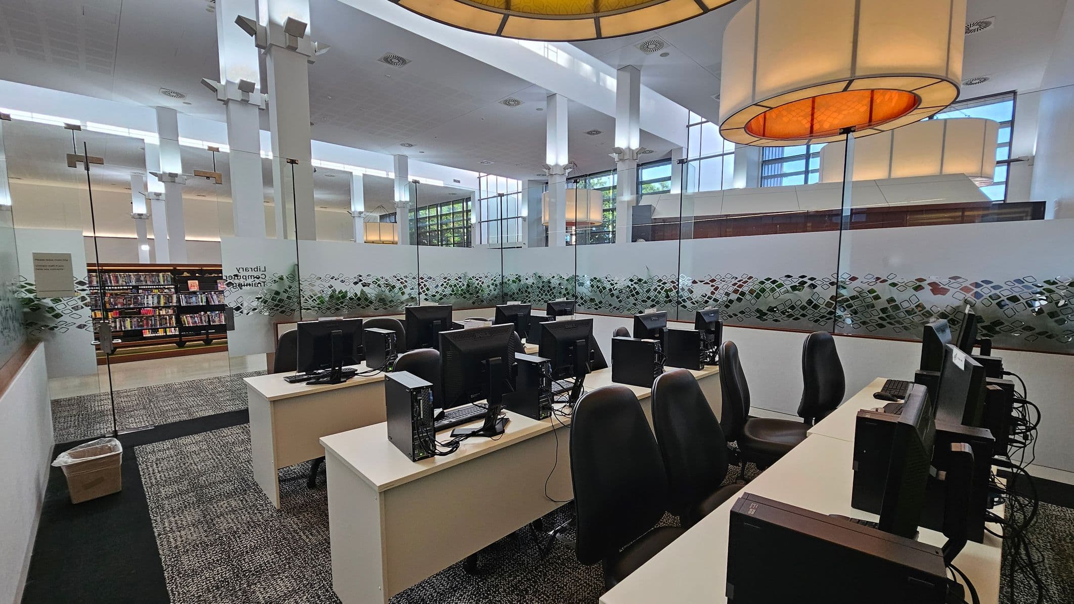 A modern library computer training room with several rows of desks, each equipped with desktop computers and black chairs. The room is enclosed by glass walls with a decorative pattern. In the background, there are bookshelves filled with books. The ceiling is high with large, circular light fixtures. The floor is carpeted with a patterned design.