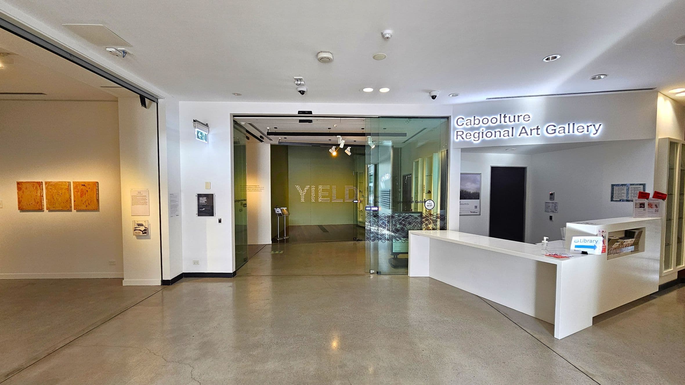 Entrance to the Caboolture Regional Art Gallery. On the left, a wall displays three square artworks with a warm colour palette. In the centre, a glass door leads into the gallery, with the word "YIELD" visible inside. On the right, a white reception desk with brochures and a hand sanitiser bottle. Above, the gallery's name is illuminated. The floor is polished concrete, and the ceiling is white with recessed lighting.