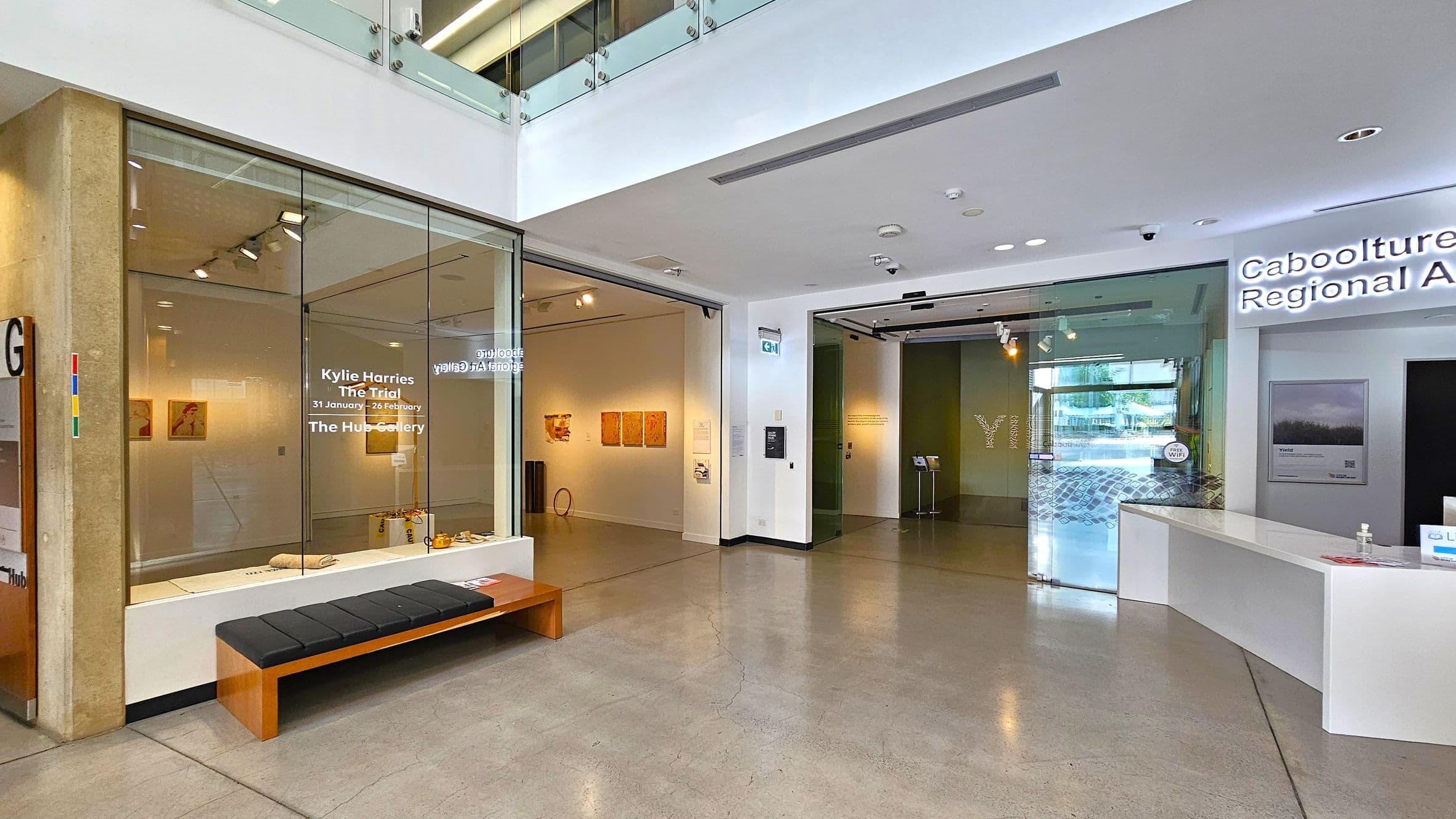 On the left, there's a glass wall with text about an exhibition by Kylie Harries at The Hub Gallery. Inside, artworks are displayed on the walls. A black cushioned bench sits in front of the glass. In the centre, an open entrance leads to more gallery space. On the right, a white reception desk is part of the Caboolture Regional Art Gallery, with a sign above. The floor is polished concrete, and the ceiling is white with recessed lighting.