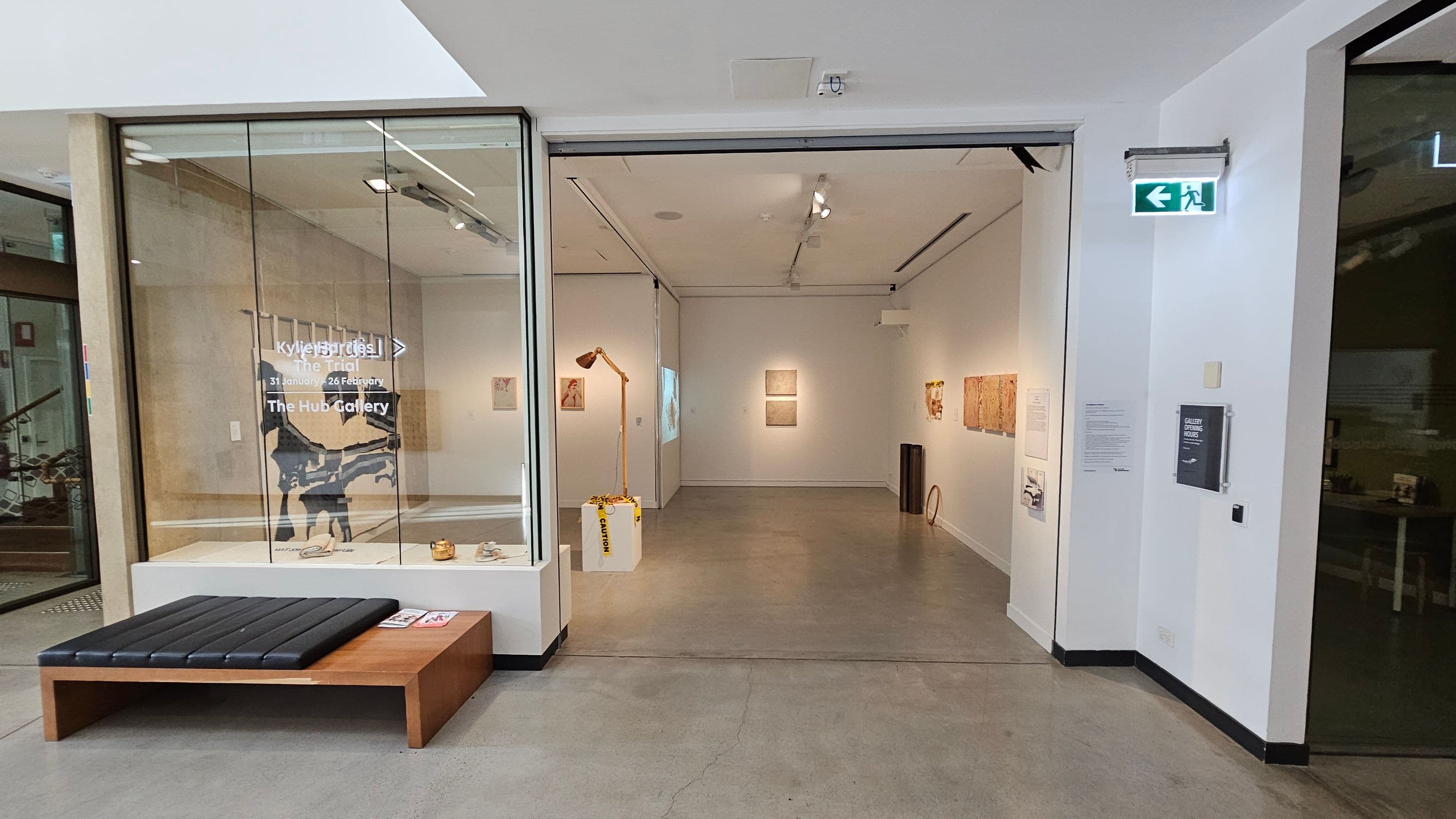 A gallery entrance with a glass wall on the left displaying text and a silhouette artwork. A wooden bench with a black cushion is in front of the glass. Inside the gallery, artworks are hung on white walls, with a yellow lamp on a white pedestal. The floor is polished concrete, and the ceiling has track lighting. An exit sign is on the right wall, along with a gallery hours sign and some text.