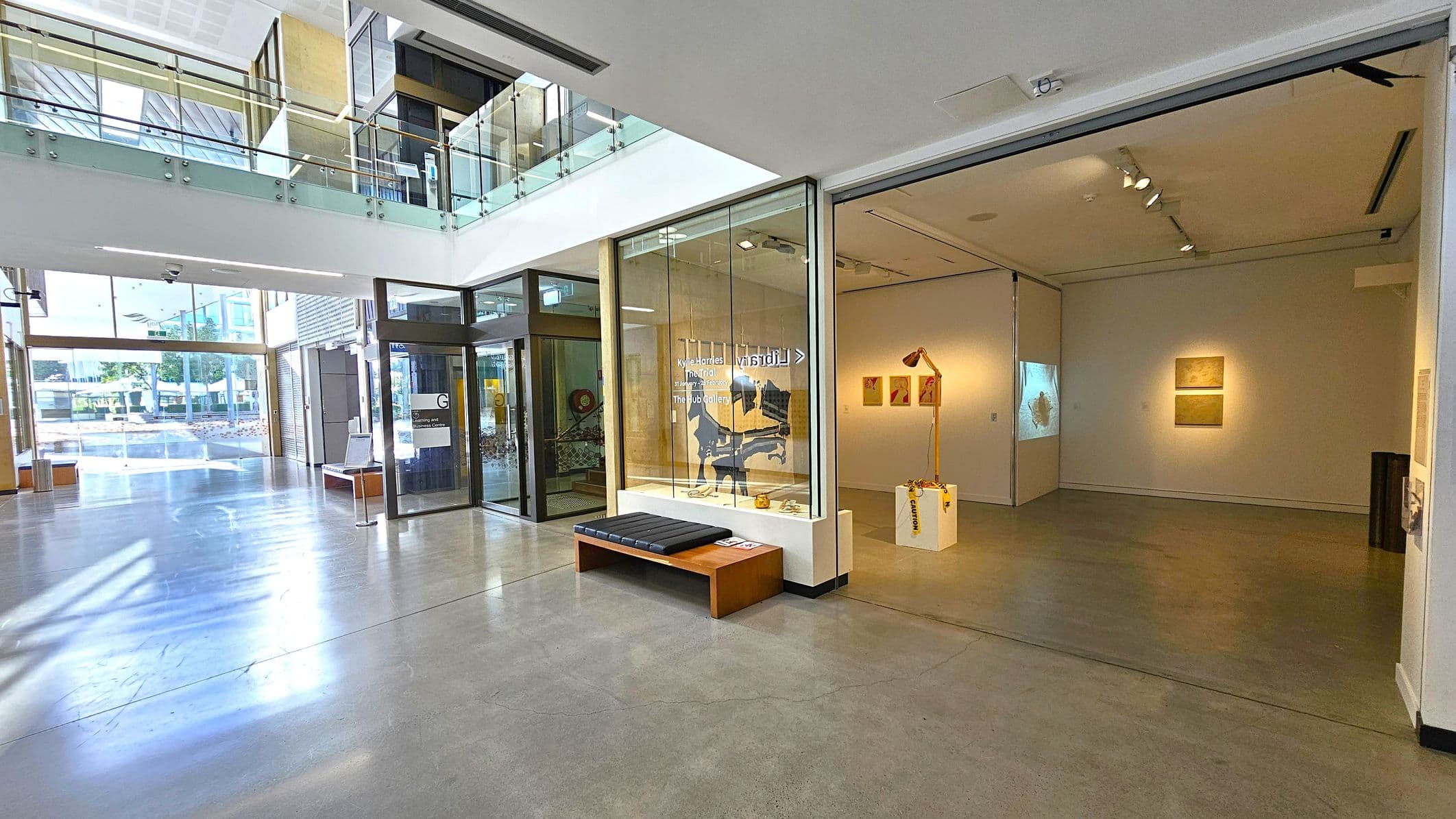 A modern interior space with a polished concrete floor. On the left, large glass doors and windows allow natural light to flood in, revealing an outdoor view. In the centre, a glass-walled area displays art, with a bench in front. To the right, an open gallery space features artworks on the walls and a sculpture on a pedestal. The ceiling is high with recessed lighting. A glass railing is visible on the upper level.