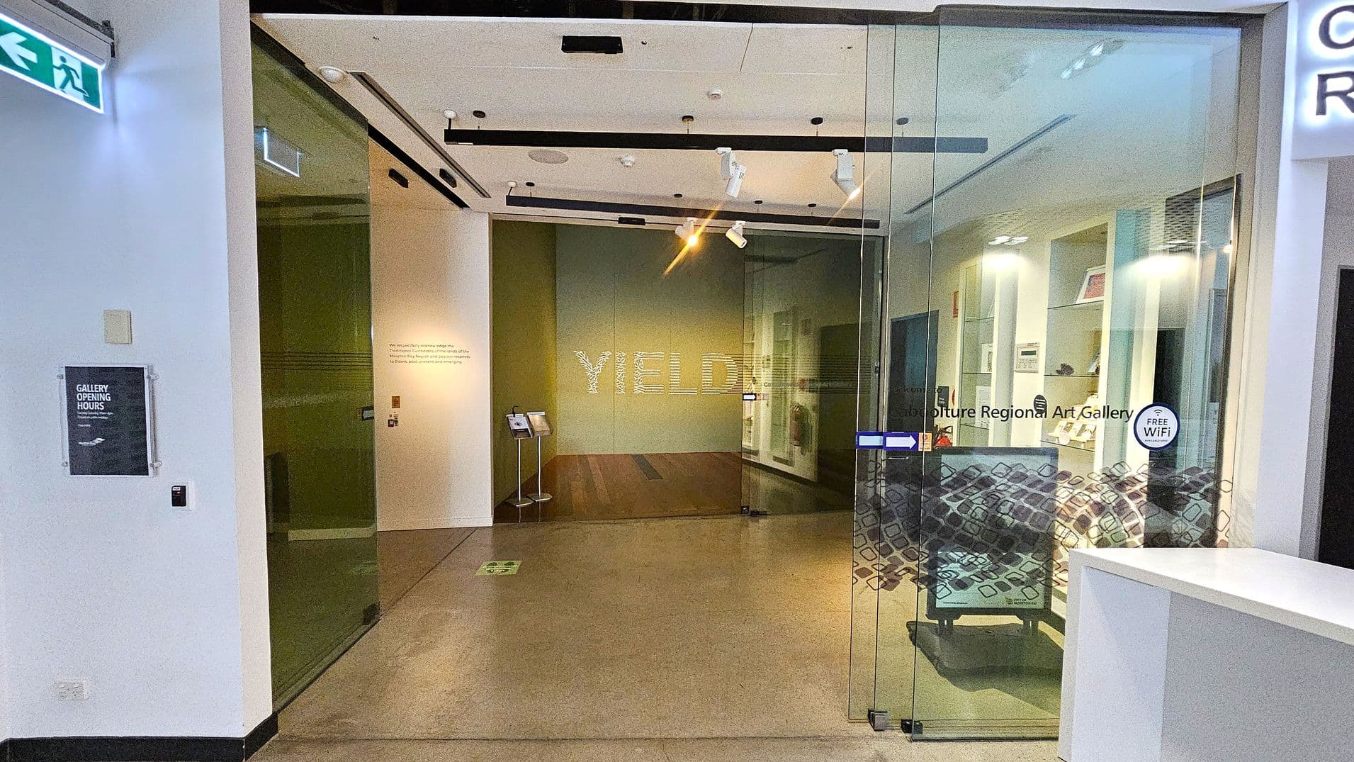 A glass entrance to a gallery with "YIELD" displayed on the back wall. On the left, a sign indicates gallery opening hours and an emergency exit sign is visible above. The right side features a glass door with "Culture Regional Art Gallery" and a "Free WiFi" sticker. Inside, there are display shelves with art pieces. The floor is a mix of polished concrete and wood, and the ceiling has track lighting.