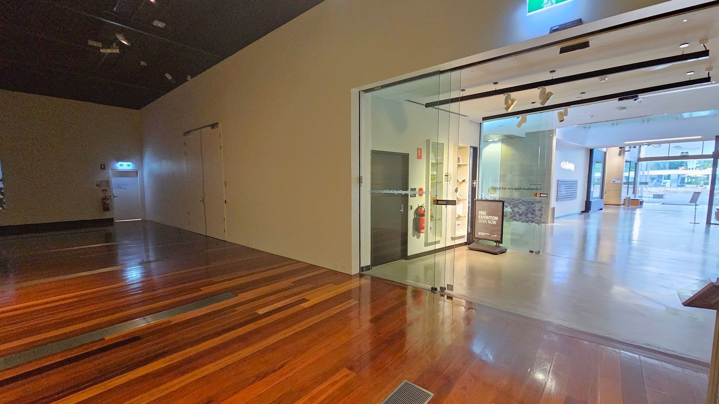 On the left, there is a dark area with polished wooden flooring and a white door with an illuminated exit sign above it. A fire extinguisher is mounted on the wall nearby. On the right, a glass entrance leads to a brightly lit space with a sign indicating a free exhibition. The floor transitions from wood to a lighter material. The ceiling is dark with spotlights, and the background shows large windows allowing natural light into the space.