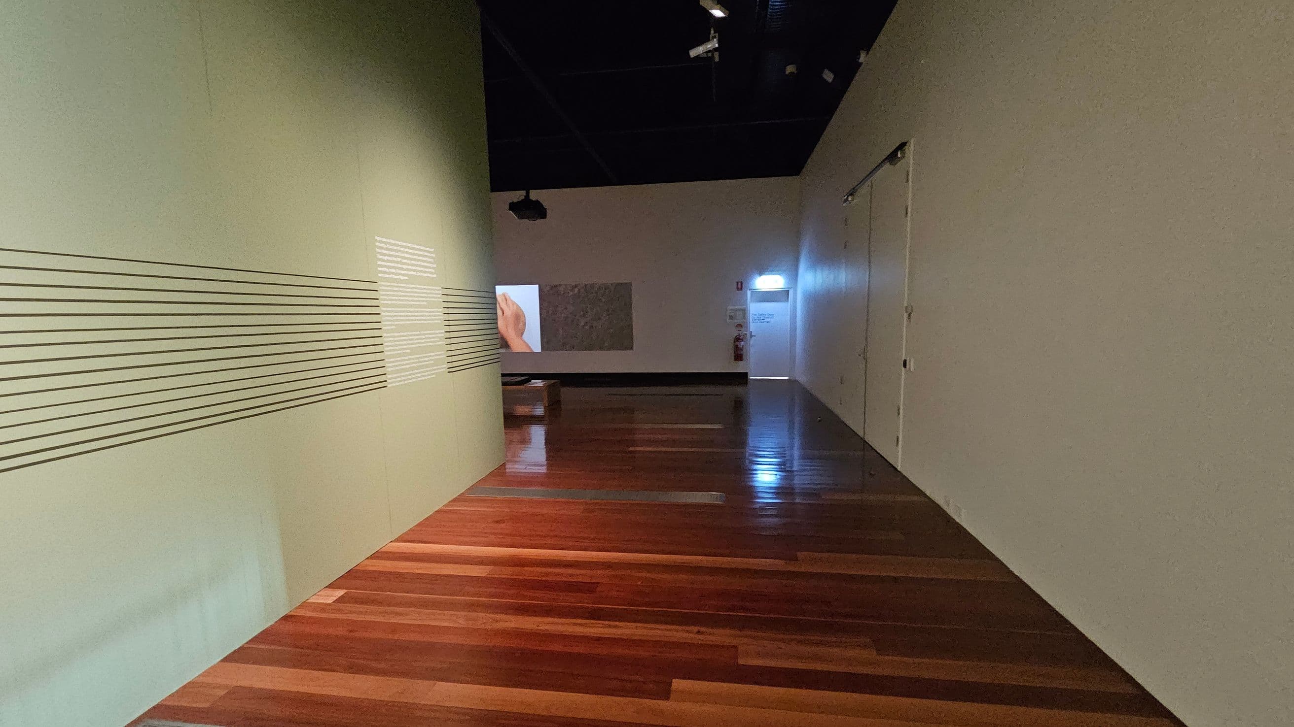 A hallway with wooden flooring and a high, dark ceiling. On the left, a green wall features horizontal black lines and a section of white text. In the background, a projection displays an image of a hand. On the right, a plain white wall with a closed door and an illuminated exit sign above another door. The space is dimly lit, creating a reflective sheen on the floor.