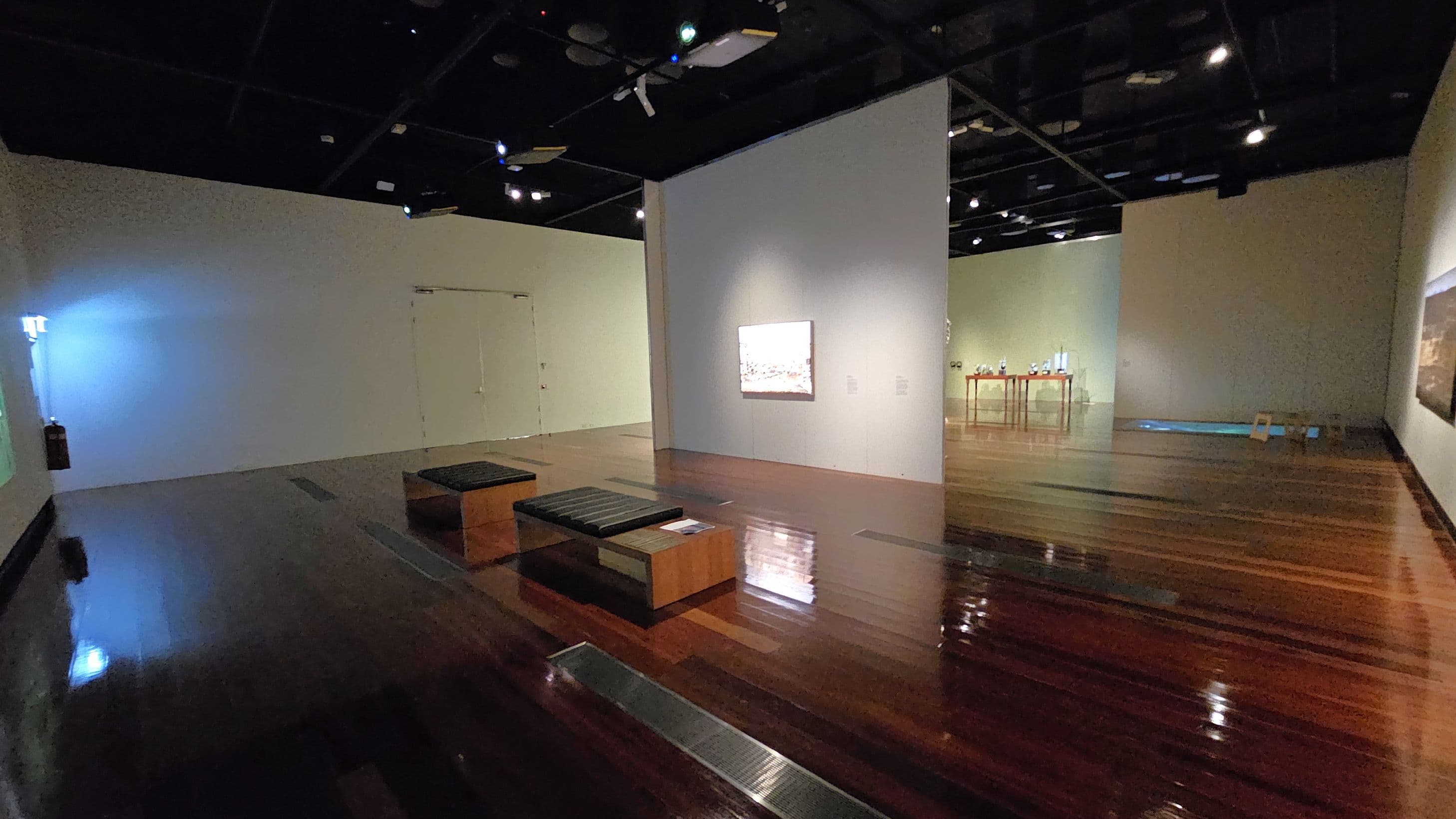 A dimly lit gallery with polished wooden floors and a dark ceiling. In the centre, a white wall displays a framed artwork. Two wooden benches with black cushions are positioned in front. To the right, a table with various objects is visible against a green wall. The left side has a plain wall with a faint blue light and a fire extinguisher. The background shows more artworks and a partially visible doorway.