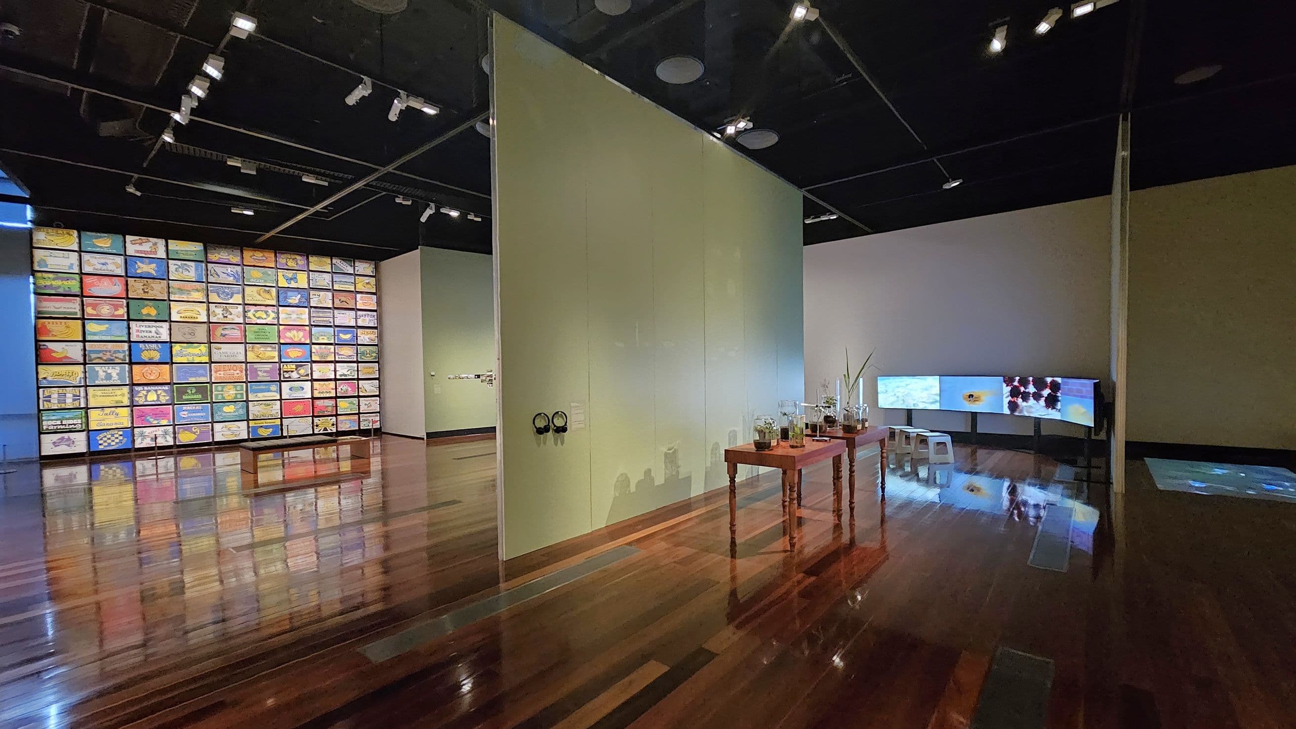A gallery space with a polished wooden floor. On the left, a large wall displays a grid of colourful, rectangular artworks. In the centre, a partition wall with headphones attached and a wooden table holding various glass containers and plants. On the right, a curved display screen shows digital images, with a small white stool in front. The ceiling is black with spotlights illuminating the exhibits.