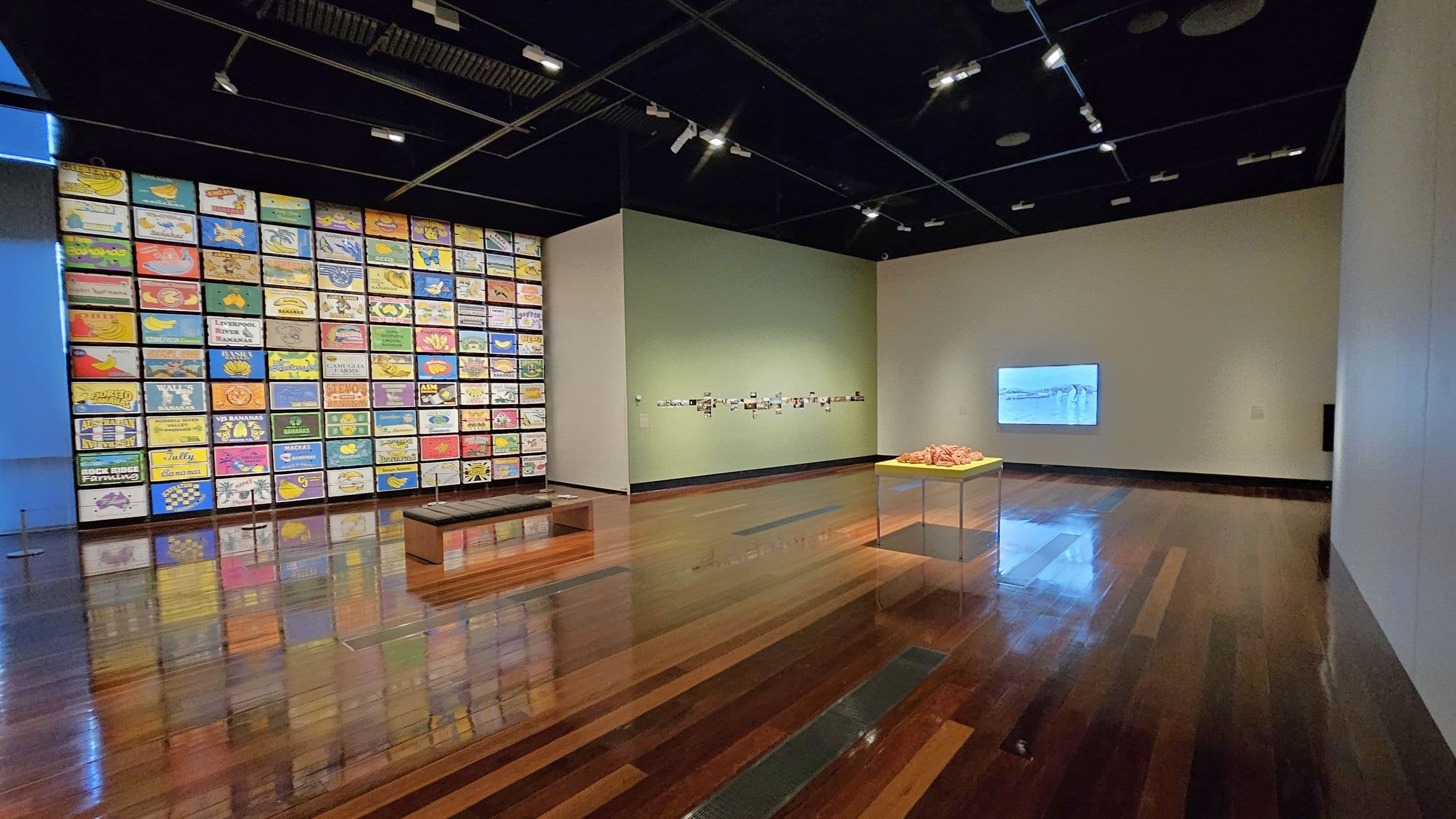 A gallery room with a polished wooden floor and a dark ceiling with spotlights. On the left wall, there is a large display of colourful rectangular artworks arranged in a grid. In the centre, a bench is placed on the floor. The right wall features a small collection of photographs and a digital screen displaying an image. In the foreground, a table with a yellow top holds a collection of objects. The walls are painted in light green and white tones.