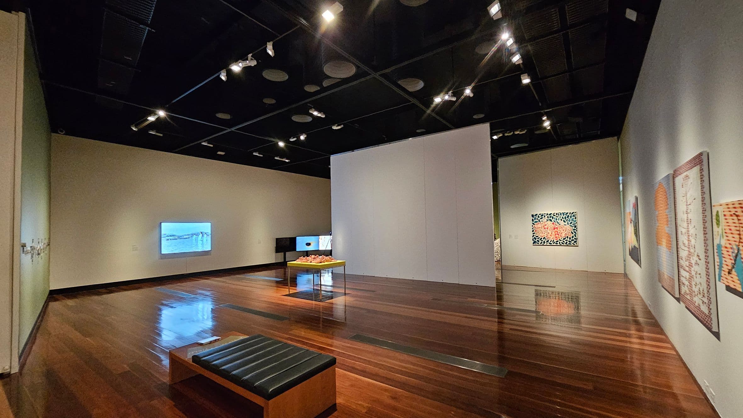 A modern art gallery with polished wooden floors and a dark ceiling with spotlights. On the left wall, a digital screen displays an image. In the centre, a table holds a sculptural piece. The right wall features several colourful artworks. A cushioned bench is positioned in the foreground. The back wall displays a vibrant painting with a patterned design.
