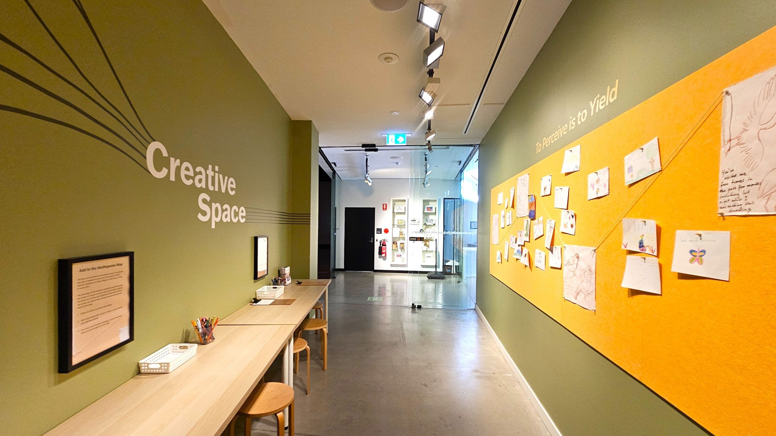 A hallway with green walls and a polished concrete floor. On the left, a long wooden table with stools, art supplies, and framed instructions. The words "Creative Space" are on the wall. On the right, a large orange board displays various drawings and notes, with the phrase "To Perceive is to Yield" above. The ceiling has track lighting, and at the end of the hallway, there's a glass door leading to another room with display cases.