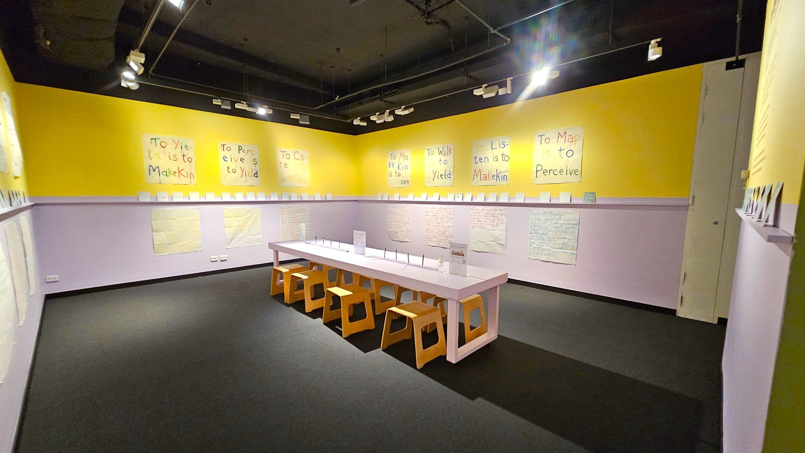 A room with yellow walls and a black ceiling, featuring handwritten posters with phrases like "To Yield is to Make Kin" and "To Map is to Perceive." A long white table with wooden stools is in the centre. The walls have a purple lower section with additional papers displayed. Track lighting is mounted on the ceiling, illuminating the space. The floor is covered with dark carpeting.