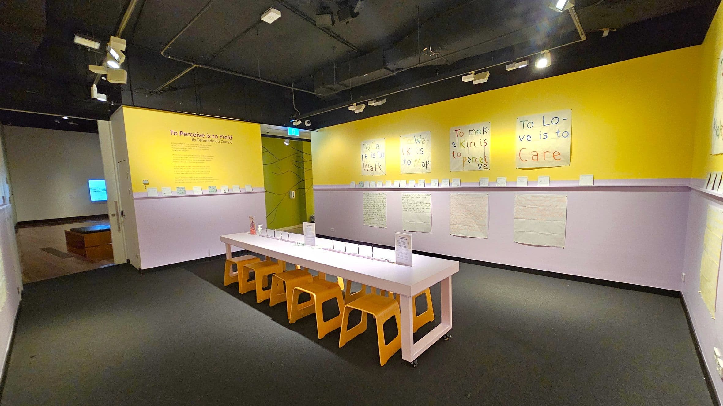 A room with yellow and lavender walls features a long white table with orange stools underneath. On the walls, there are handwritten posters with phrases like "To Love is to Care" and "To make Kin is to perceive." Above the posters, smaller framed items are displayed on a ledge. The ceiling is black with exposed lighting fixtures. In the background, a doorway leads to another room with a bench and a screen. The floor is carpeted in dark grey.