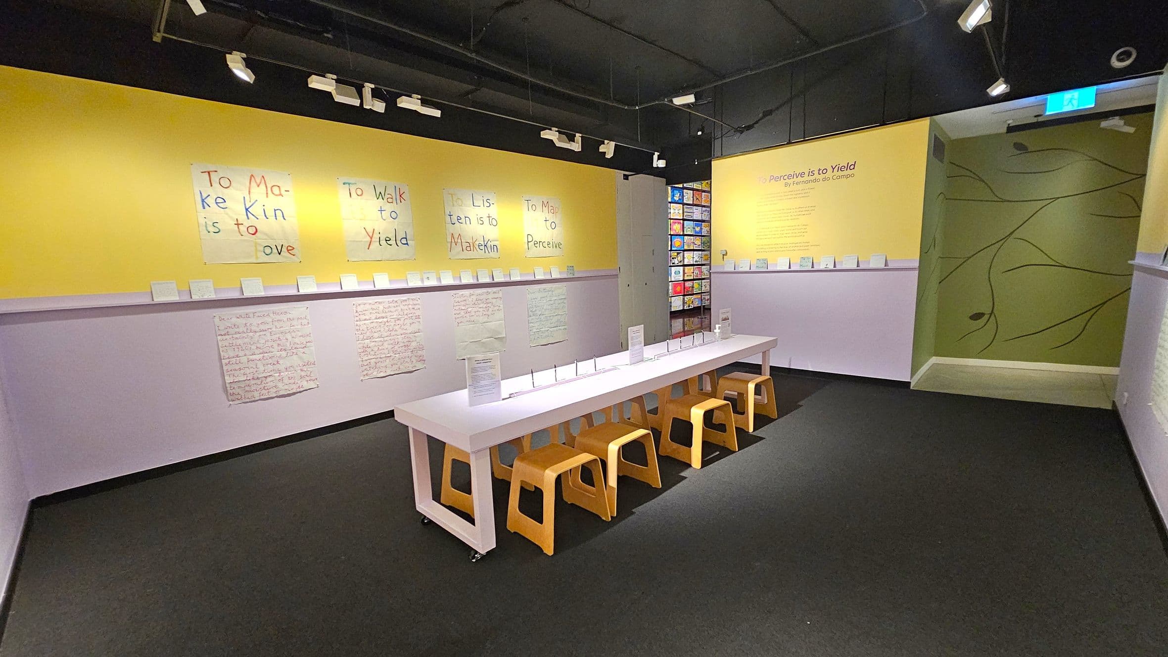 A room with a black ceiling and track lighting. The left wall is yellow with handwritten posters displaying phrases like "To Make Kin is to Love." Below, a purple shelf holds small cards. A long white table with wooden stools is in the centre. The right wall is yellow with text, leading to a green wall with abstract lines. The floor is dark grey carpet.