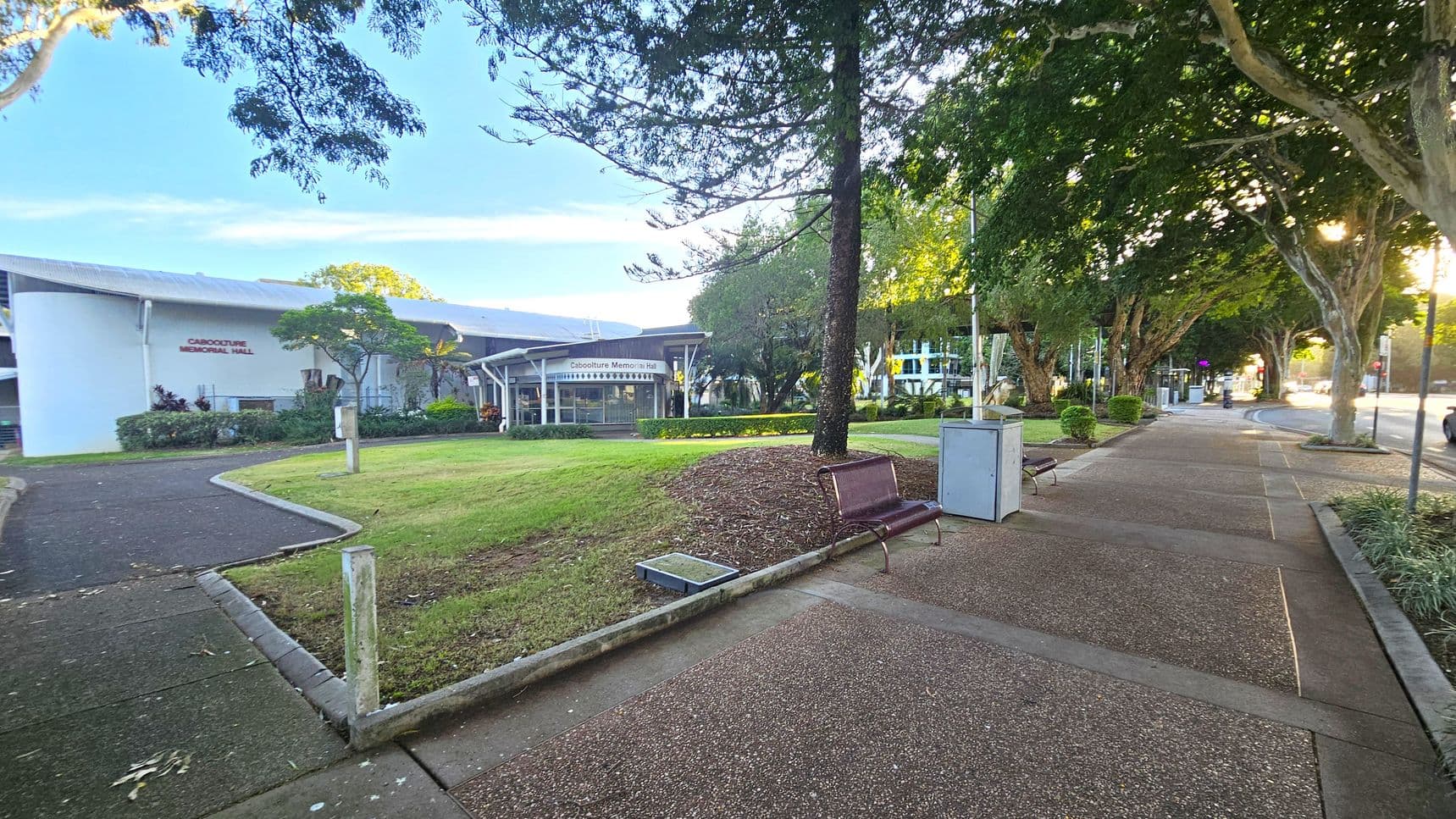 A paved footpath runs along the right side, bordered by trees and a grassy area. On the left, a white building with a sign reading "Caboolture Memorial Hall" is visible. In the foreground, there is a bench and a bin near the path. The background features more trees and a road extending into the distance. The area is well-maintained with trimmed grass and landscaped plants.