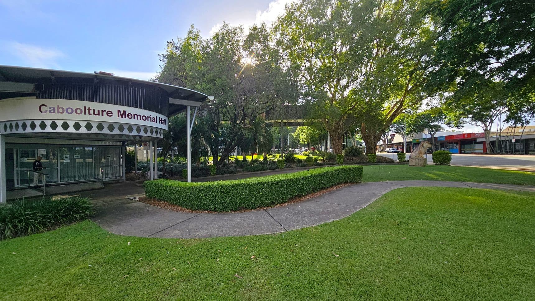 Caboolture Memorial Hall is on the left with a curved roof and glass entrance. A person is walking near the entrance. In the centre, there are neatly trimmed hedges and a path leading through a garden with trees. On the right, a street with shops and a stone sculpture of a lion is visible. The area is surrounded by green grass and trees, with sunlight filtering through the leaves.