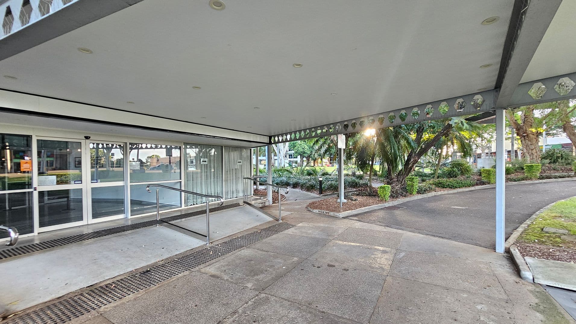A covered walkway with a grey ceiling featuring recessed lights and decorative cut-outs. On the left, there are glass sliding doors with a ramp and metal handrails leading up to them. The floor is concrete with drainage grates. On the right, a path curves through a landscaped garden with palm trees and shrubs. Sunlight filters through the trees, creating a bright spot in the greenery.