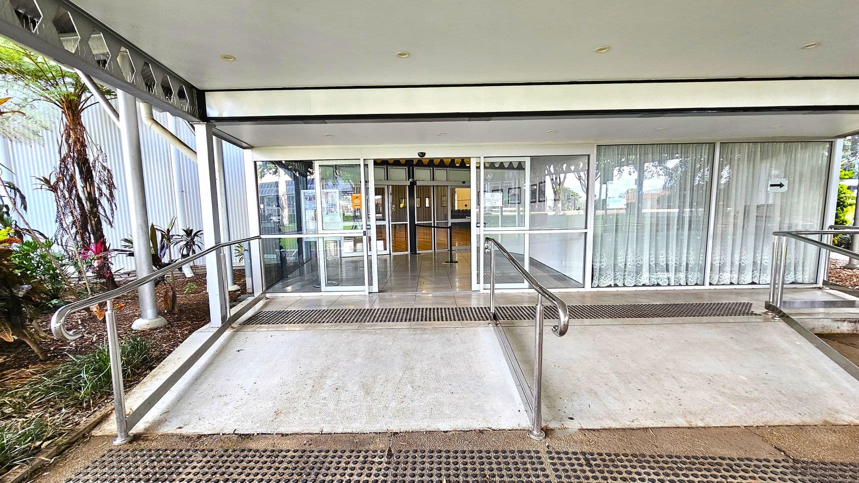 A concrete ramp with metal railings leads to a glass entrance with sliding doors. The doors are flanked by large windows with sheer curtains. On the left, there are plants and trees in a garden bed. The ceiling is white with recessed lights, and the floor is tiled near the entrance. The area is covered, providing shelter to the entrance.