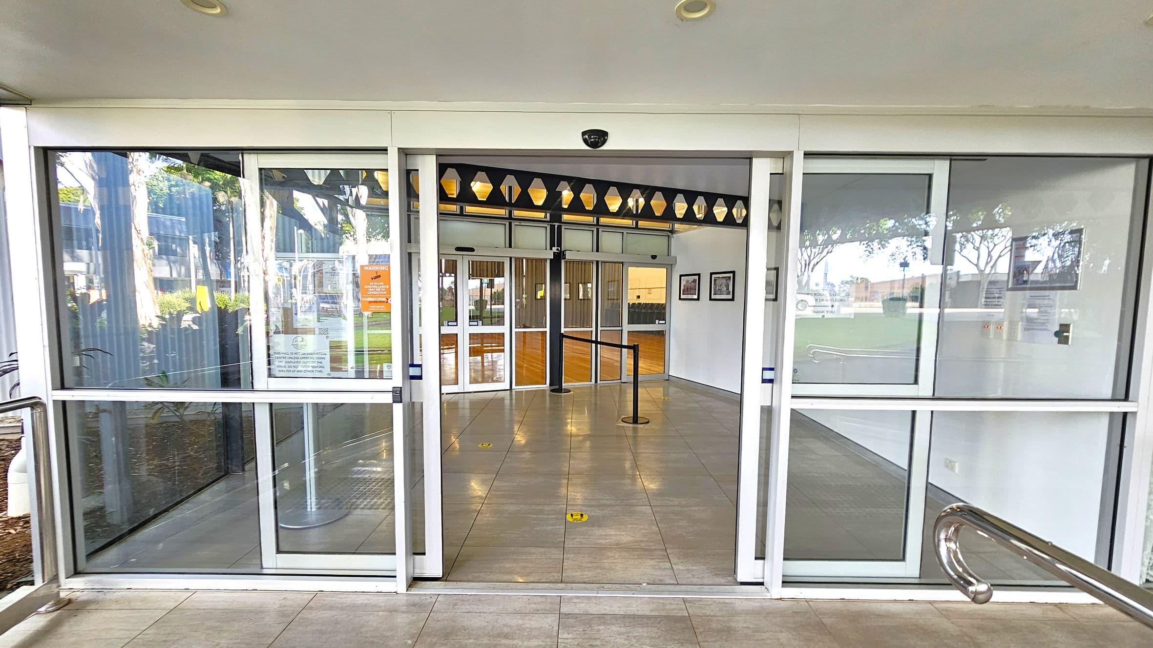 Automatic sliding glass doors lead into a building. The floor is tiled, and there is a metal railing on the right. Inside, a wooden floor is visible, along with a queue barrier. The ceiling is white with a security camera. On the left, there are windows reflecting trees and a building. On the right, framed pictures hang on the wall. Signs and notices are posted on the glass doors.