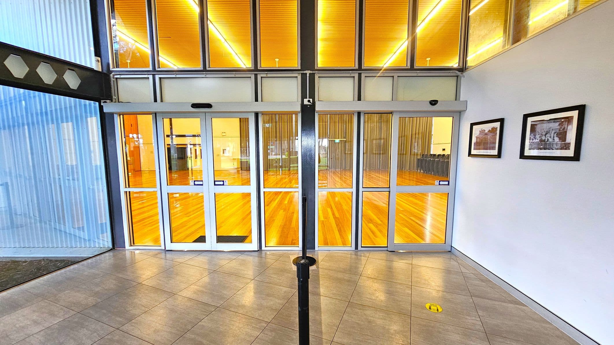 A hallway with a tiled floor leads to a set of glass double doors in the centre, revealing a room with a wooden floor and bright lighting. On the right wall, two framed pictures are displayed. The left side features a glass wall with vertical blinds. Above the doors, there are additional glass panels with a warm glow from the ceiling lights.