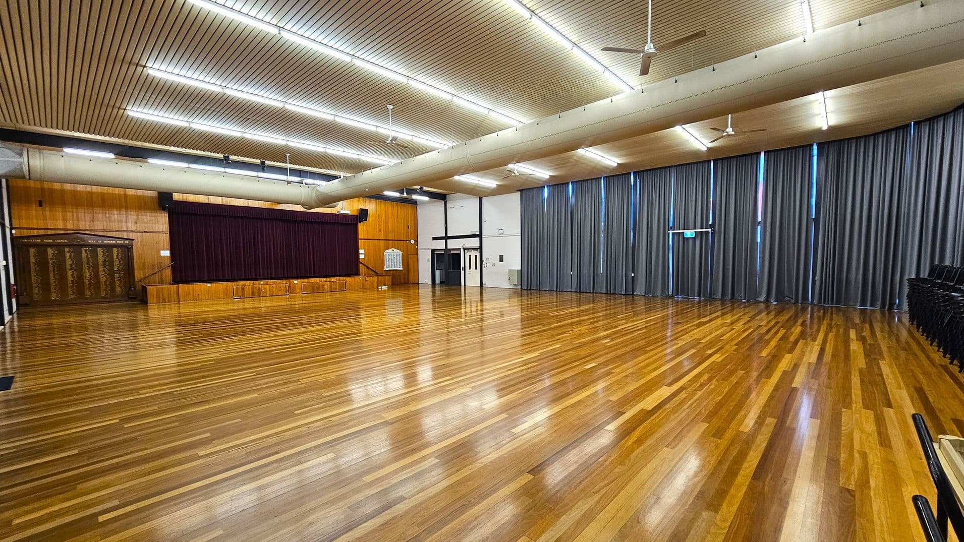 A spacious hall with polished wooden flooring. On the left, there's a stage with a maroon curtain and wooden panelling. The right side features tall grey curtains covering windows, with a stack of black chairs in the corner. The ceiling is lined with fluorescent lights and has a large ventilation duct running across it.