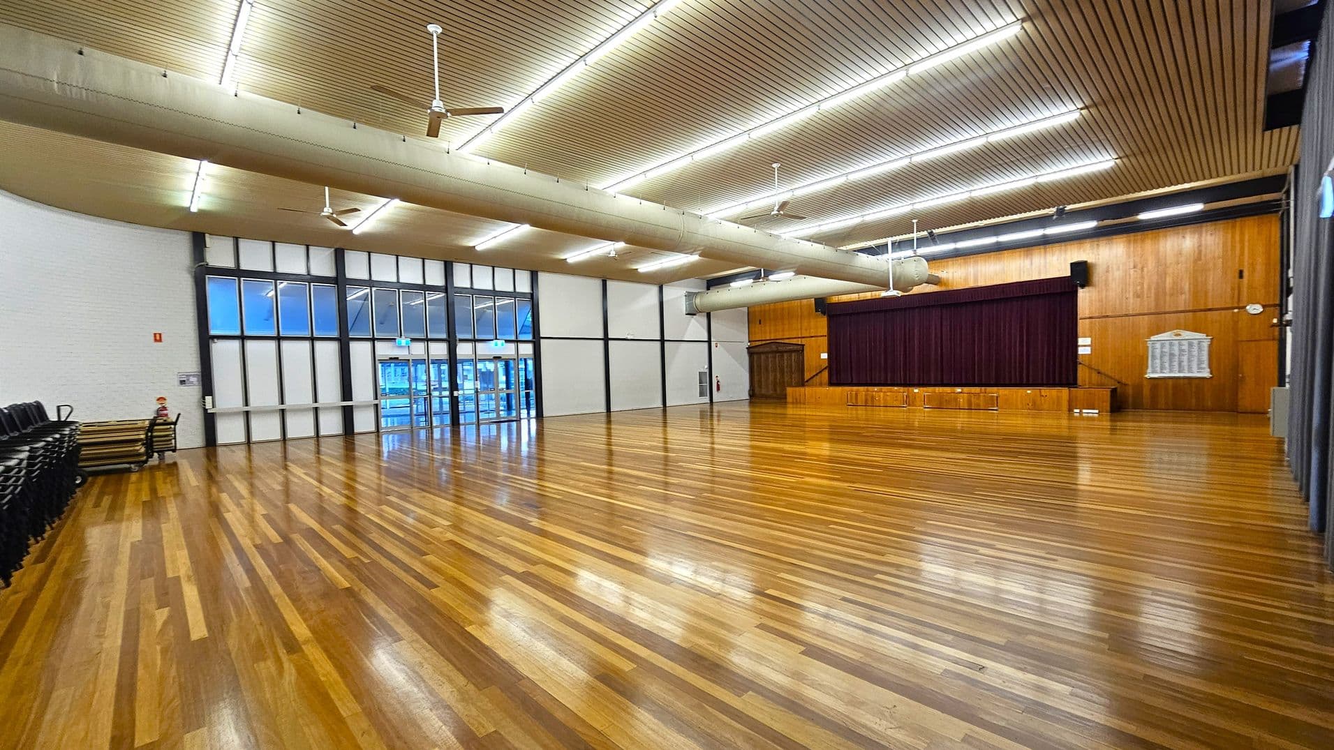 A spacious hall with polished wooden flooring. On the right, there's a stage with a maroon curtain and wooden panelling. The left side has stacked chairs and tables against a white wall. The back wall features large windows and doors with a grid design. The ceiling is lined with wooden slats and equipped with fluorescent lights and ceiling fans. A large air duct runs across the ceiling.