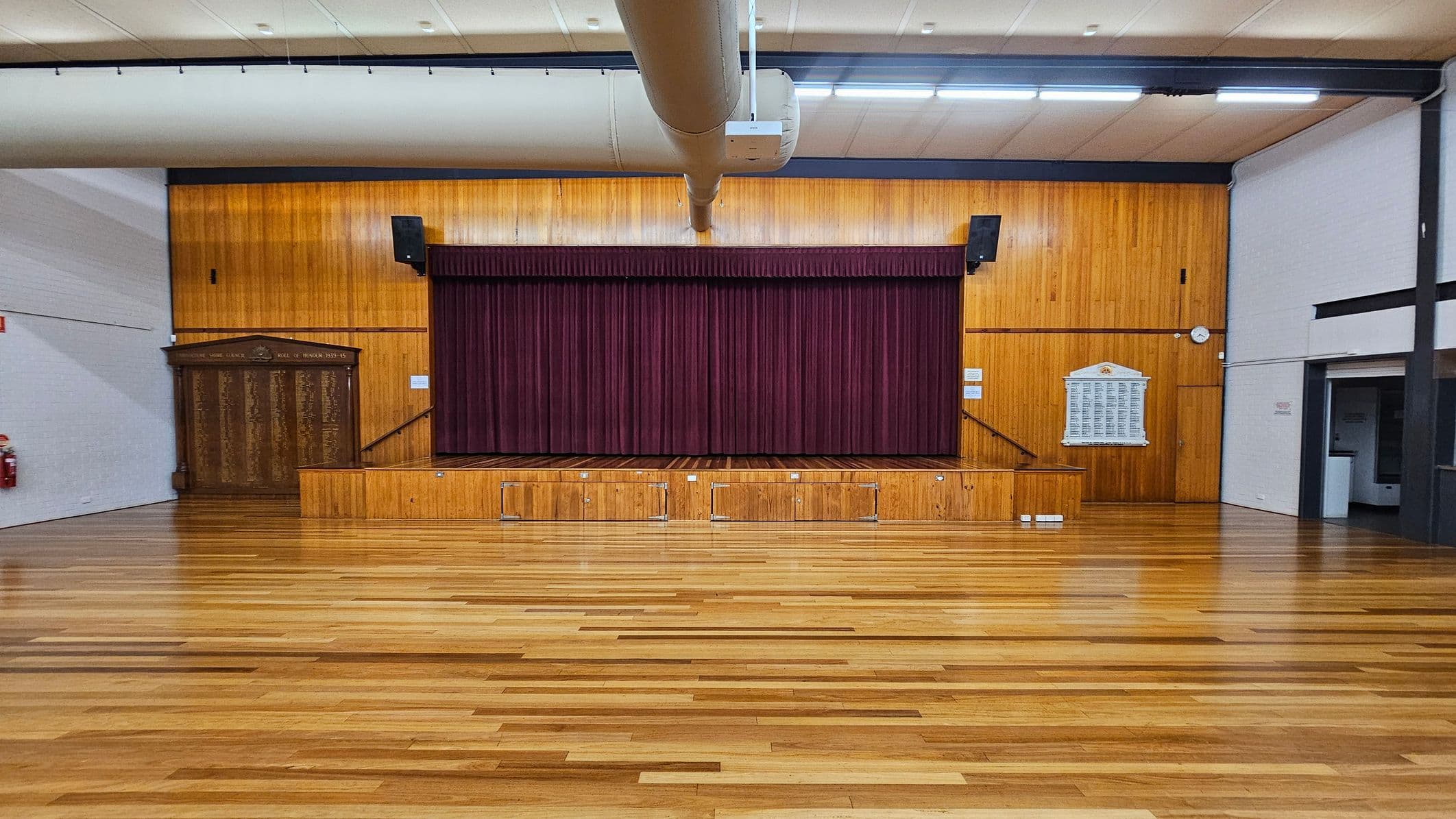 A large hall with a polished wooden floor and a stage at the centre. The stage has a maroon curtain and is flanked by wooden panelling. On the left, there's a wooden plaque and a fire extinguisher. On the right, a white plaque is mounted on the wall, and a clock is visible. The ceiling has a large duct running across it with fluorescent lights. The walls are a mix of wood and white surfaces.