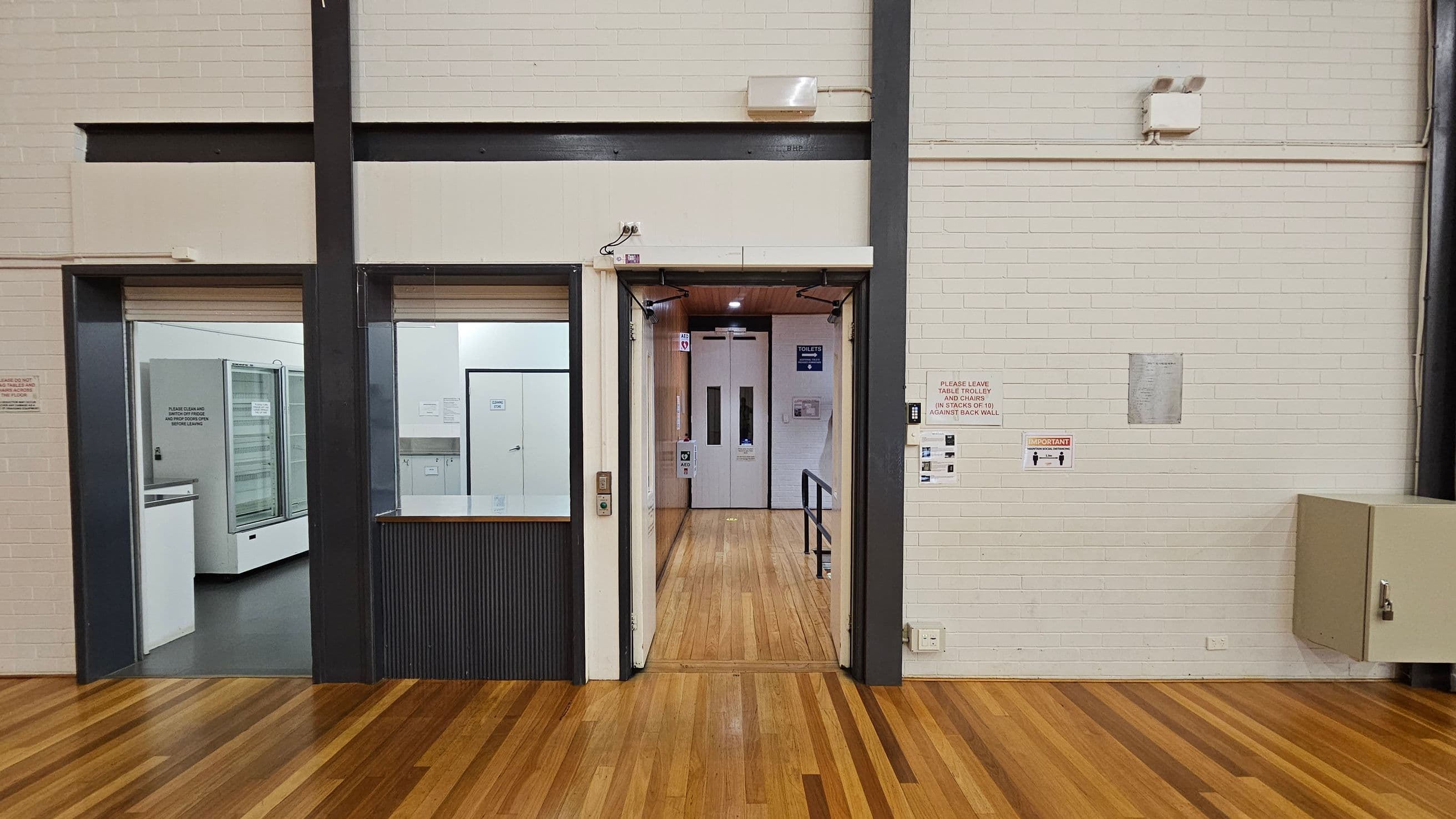 A room with light-coloured brick walls and a wooden floor. In the centre, there is an open doorway leading to a corridor with a sign indicating toilets. On the left, a room with glass-fronted refrigerators is visible through a large window. On the right, there are several signs on the wall, including one about stacking tables and chairs. A beige cabinet is mounted on the wall to the right. The ceiling is not visible.