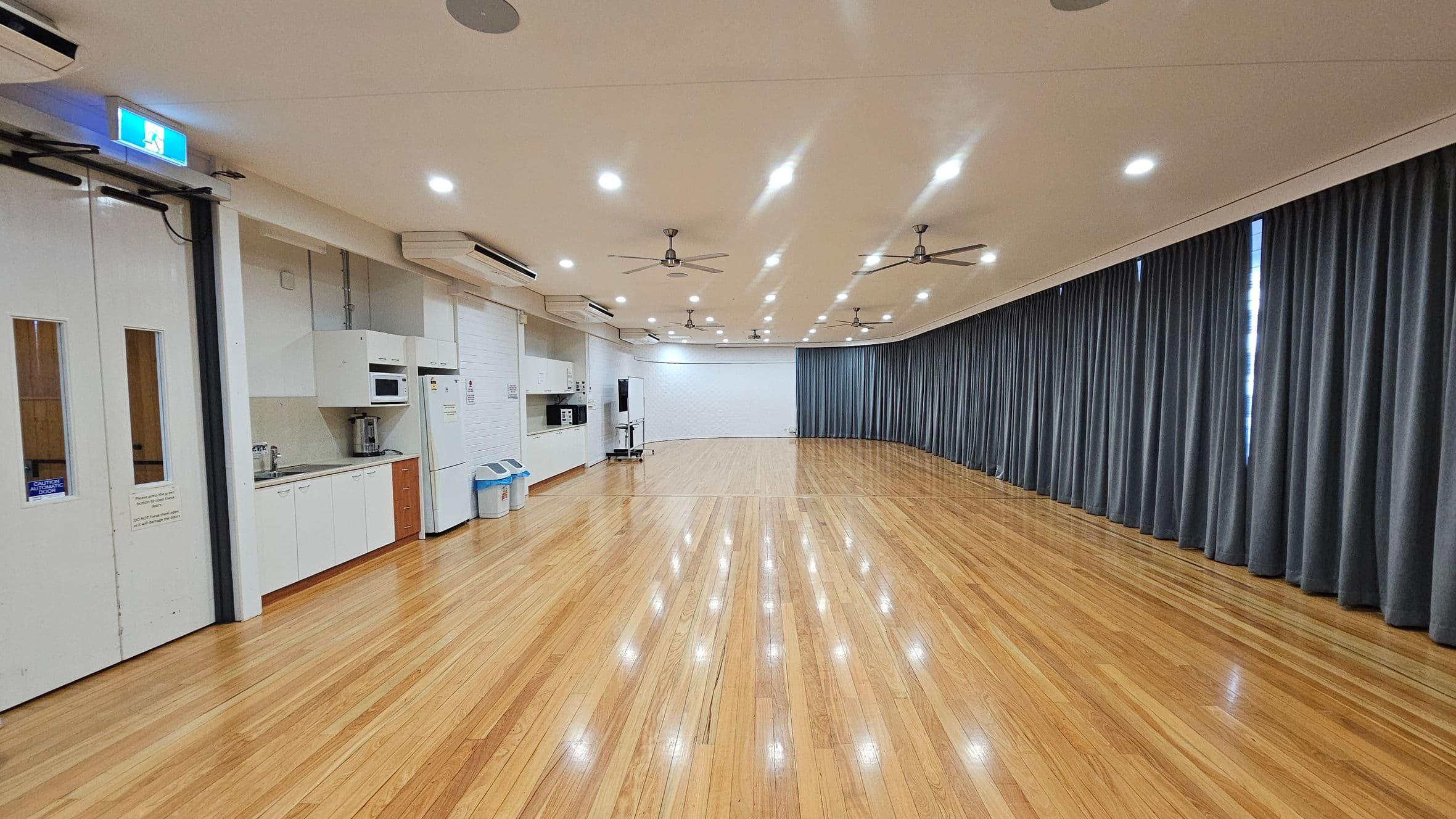 A spacious room with polished wooden floors and a high ceiling featuring multiple ceiling fans and recessed lights. On the left, there is a kitchenette with white cabinets, a microwave, a fridge, and a sink. Two bins are placed nearby. The right side is lined with long, dark curtains covering windows. At the far end, a whiteboard is visible. The room has a clean, modern appearance.
