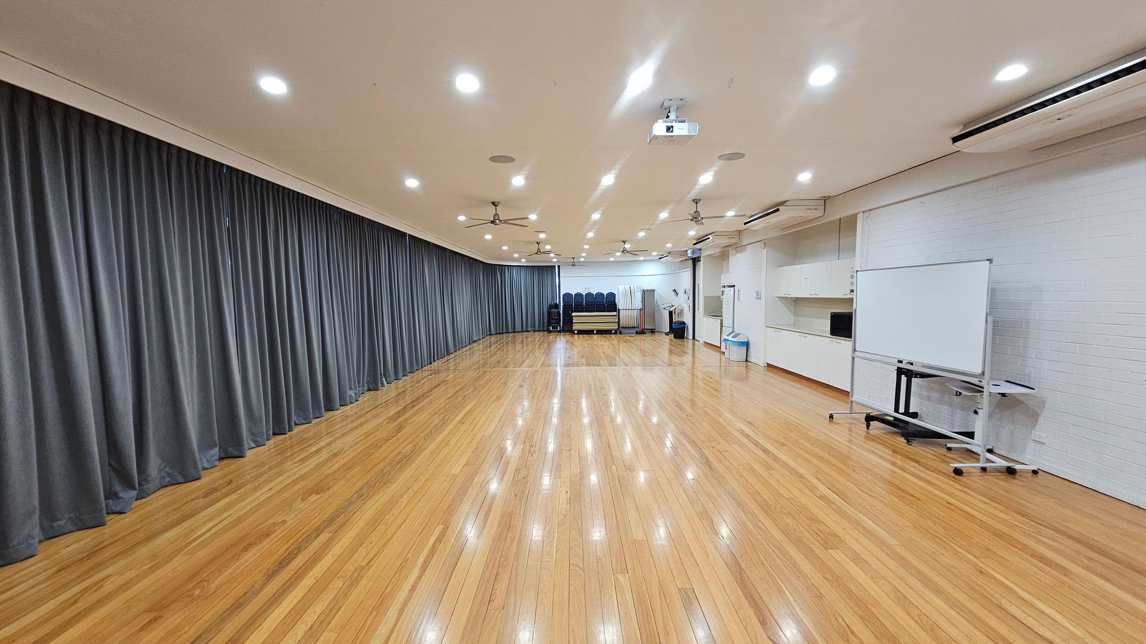 A spacious room with polished wooden floors and a ceiling fitted with recessed lights and ceiling fans. On the left, there are long grey curtains covering the wall. The right side features a whiteboard on wheels and a white brick wall with shelves and a bin. At the far end, there are stacked chairs and tables. A projector is mounted on the ceiling.
