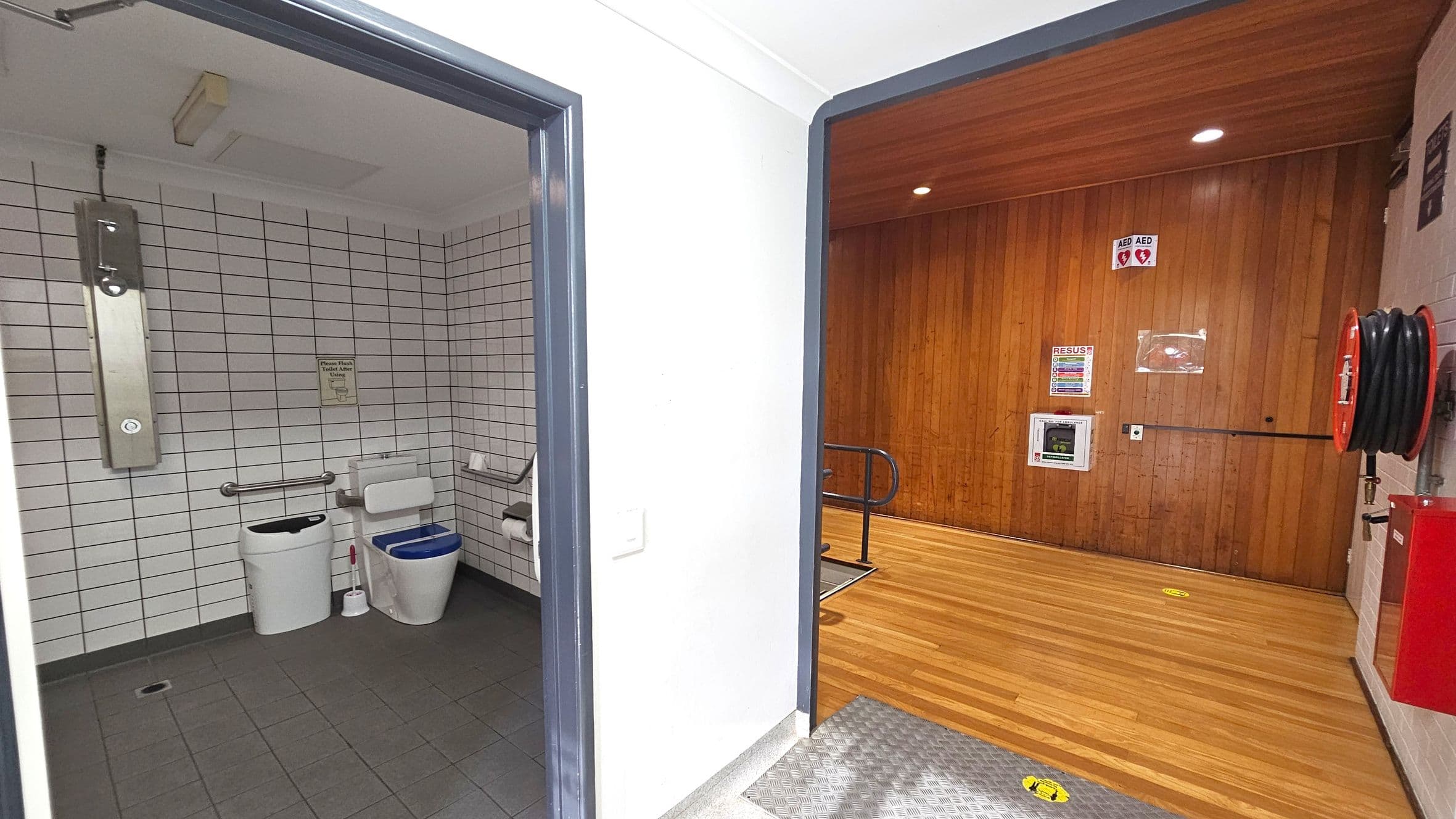 On the left, a public toilet with white tiled walls, a toilet with a blue seat, a waste bin, and a toilet brush. There are grab bars and a toilet paper holder. On the right, a room with wooden panelled walls and a wooden floor. A fire hose reel is mounted on the wall, along with an AED sign and a resuscitation guide. The ceiling is white with recessed lighting.