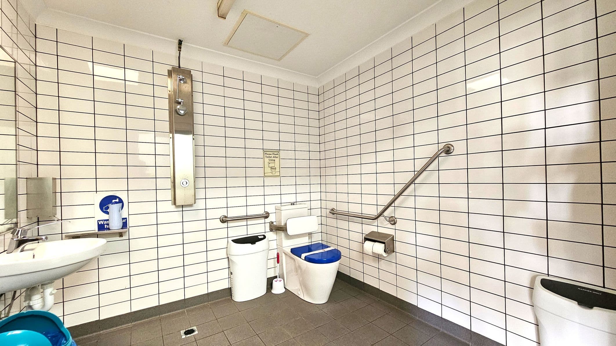 A public restroom with white tiled walls and a grey tiled floor. On the left, there's a sink with a soap dispenser and a mirror above it. In the centre, a toilet with a blue seat cover is equipped with support rails on both sides. A waste bin and a toilet brush are nearby. On the right, a toilet paper holder is mounted on the wall. The ceiling is white with a light fixture.