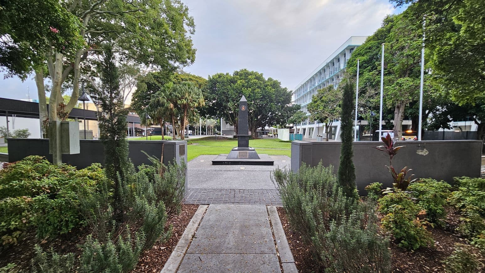 A pathway leads to a black obelisk monument in the centre, surrounded by a well-maintained garden with various shrubs and small trees. On the left, there are large trees and a building with a dark roof. On the right, a modern white building with multiple windows is visible, along with several flagpoles. The background features more trees, creating a lush, green environment. The ground is paved with concrete and gravel.