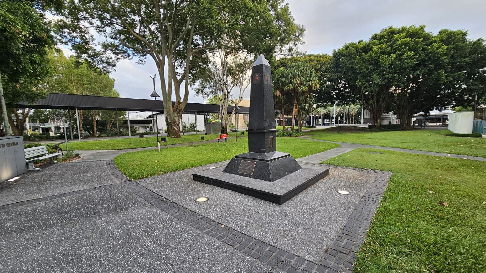 A black obelisk monument stands on a paved area with a plaque at its base. Surrounding it is a grassy park with pathways leading in different directions. To the left, there's a white bench and a low wall with text. In the background, tall trees provide shade, and a long, dark canopy structure stretches across the park. A red bench is visible near the centre, and buildings can be seen in the distance.